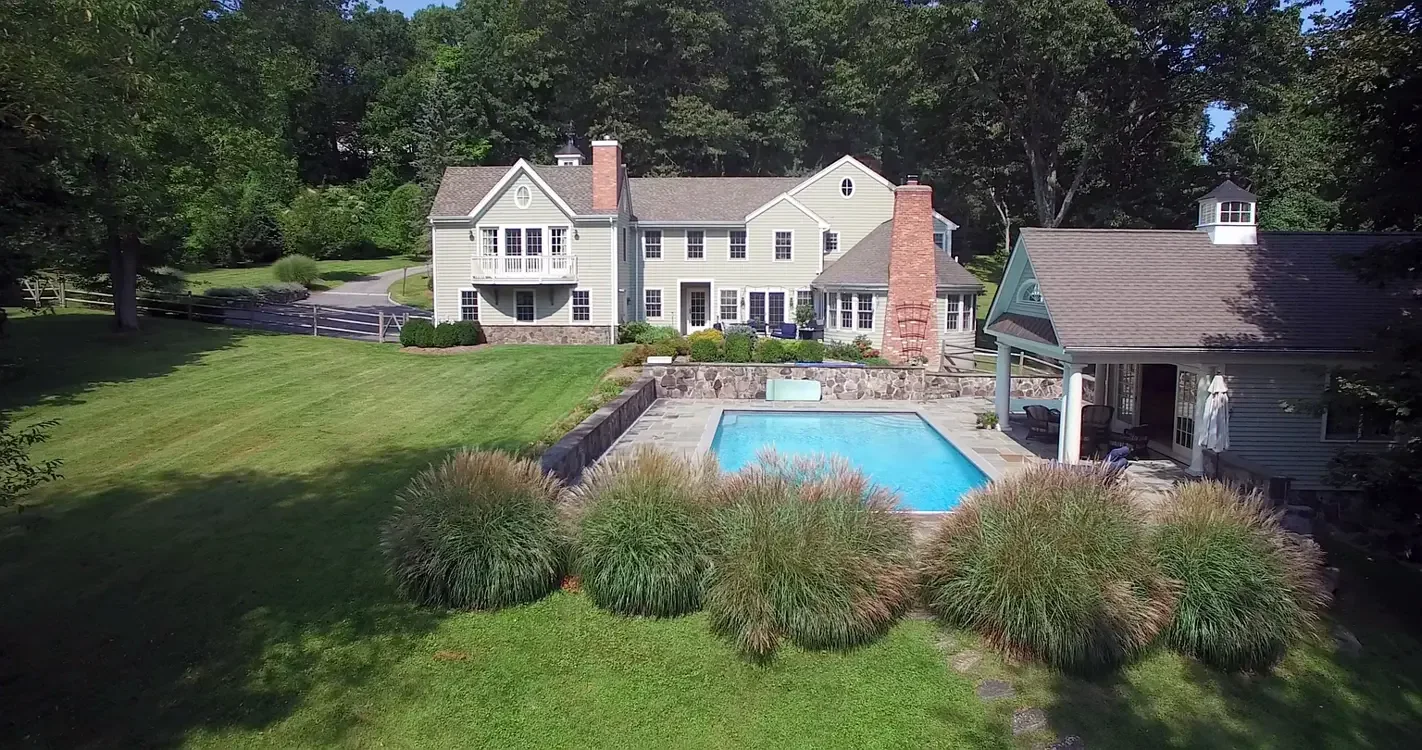 Large backyard with a rectangular swimming pool, surrounded by stone patio and lush green lawn. There is a two-story house with a white exterior, multiple windows, a balcony, and a chimney, along with a smaller structure with a covered patio area.