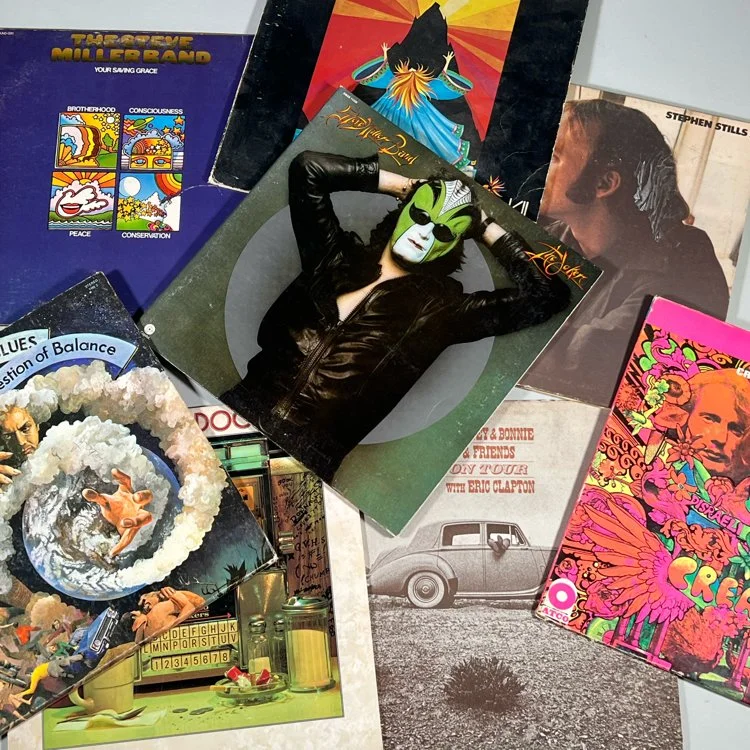 Collection of vinyl record album covers featuring various artwork and photographs, including a person with a green mask and black leather jacket lying on one cover, and a colorful floral design with pink, green, and orange accents, auctioned and phot