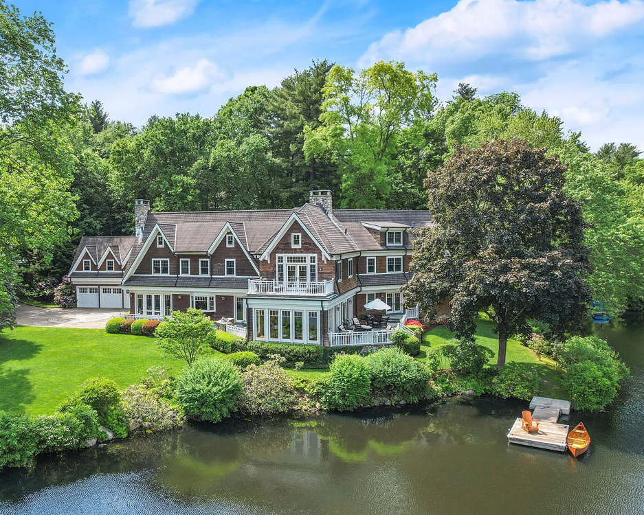 Large house surrounded by lush green trees and lawn, located near a small body of water with a dock and a boat, under a partly cloudy blue sky.