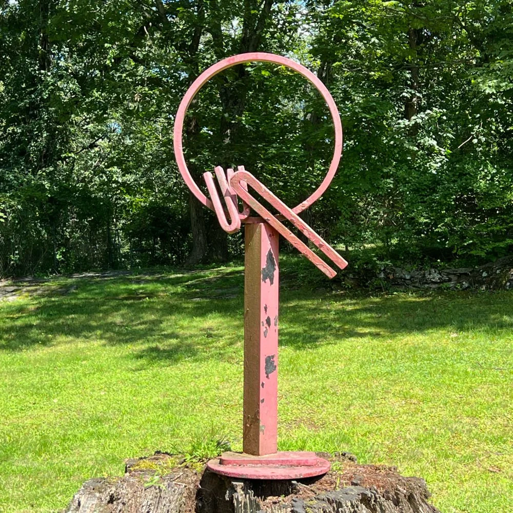 A weathered pink metal sculpture resembling a pair of eyeglasses, with the word "UK" hanging from the upper part of the frame, positioned on a wooden stump in a grassy area with trees in the background, auctioned and photographed by ACES Gallery.