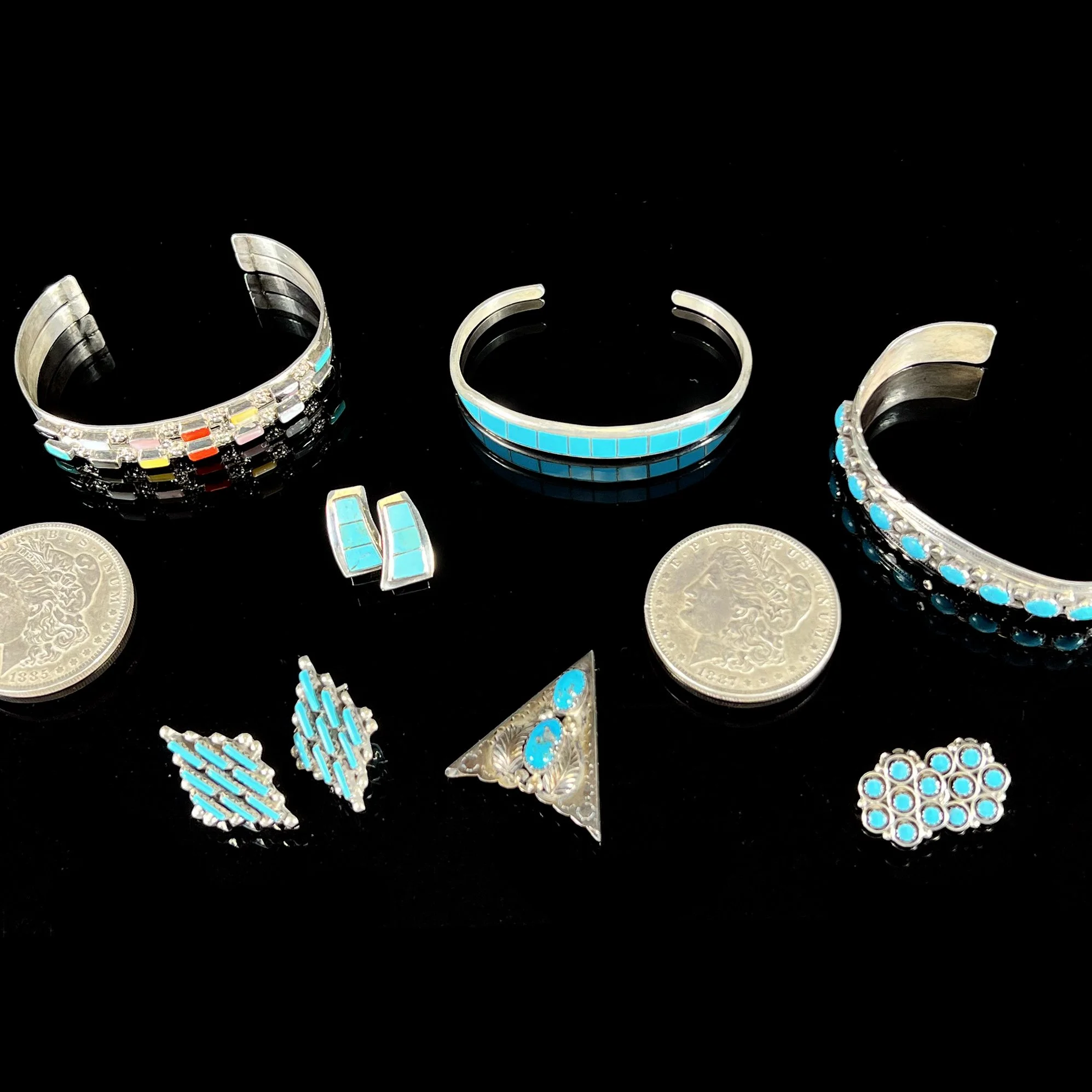 Silver jewelry including three cuff bracelets and five rings, all with turquoise and colorful accents, arranged around two coins on a black background, auctioned and photographed by ACES Gallery.