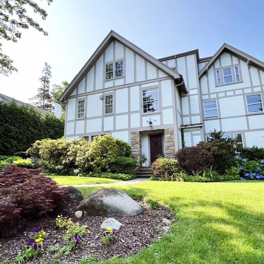 A large, white, Tudor-style house with dark timber accents, a stone front porch, and trimmed shrubs and colorful flowers in the front yard, photographed and represented by ACES.