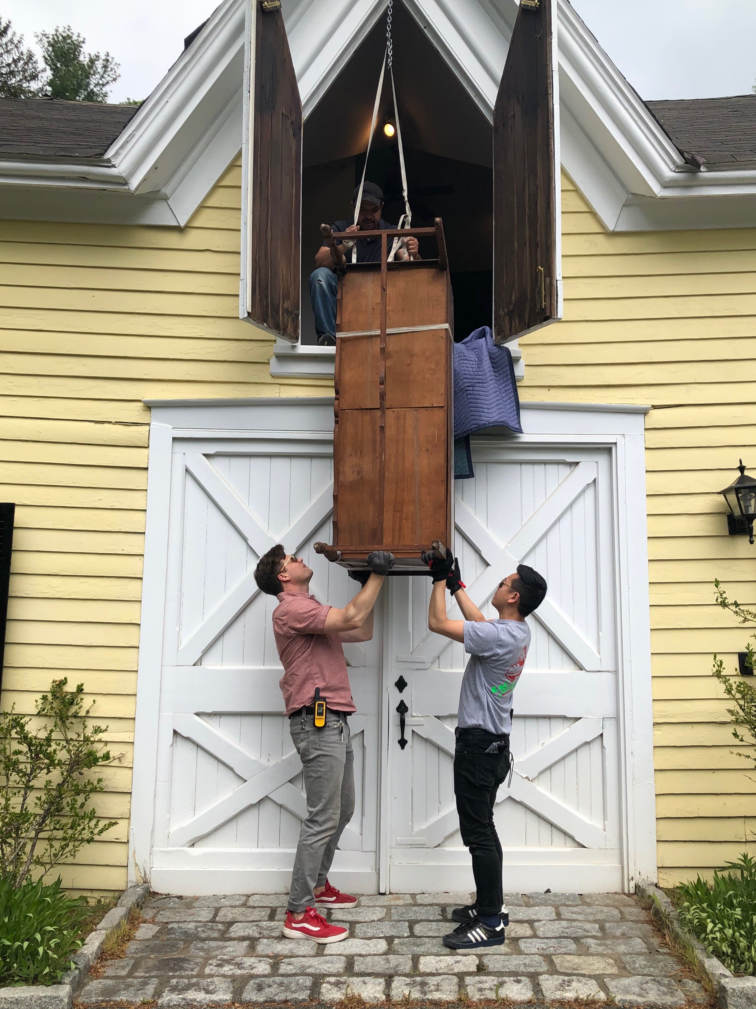 Two men are lifting a wooden piece of furniture into a second-floor window of a yellow house with white double doors and shutters. Inside, another person is standing behind the window, guiding the furniture, performed & photographed by ACES Gallery.