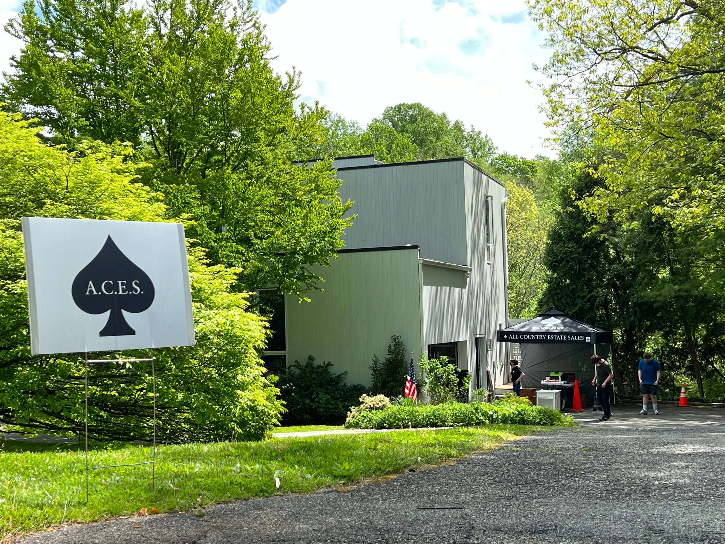 A sign with A.C.E.S. and a spade symbol, a small outdoor sales booth with a canopy reading 'All Country Estate Sales,' and people browsing in a lush green setting.