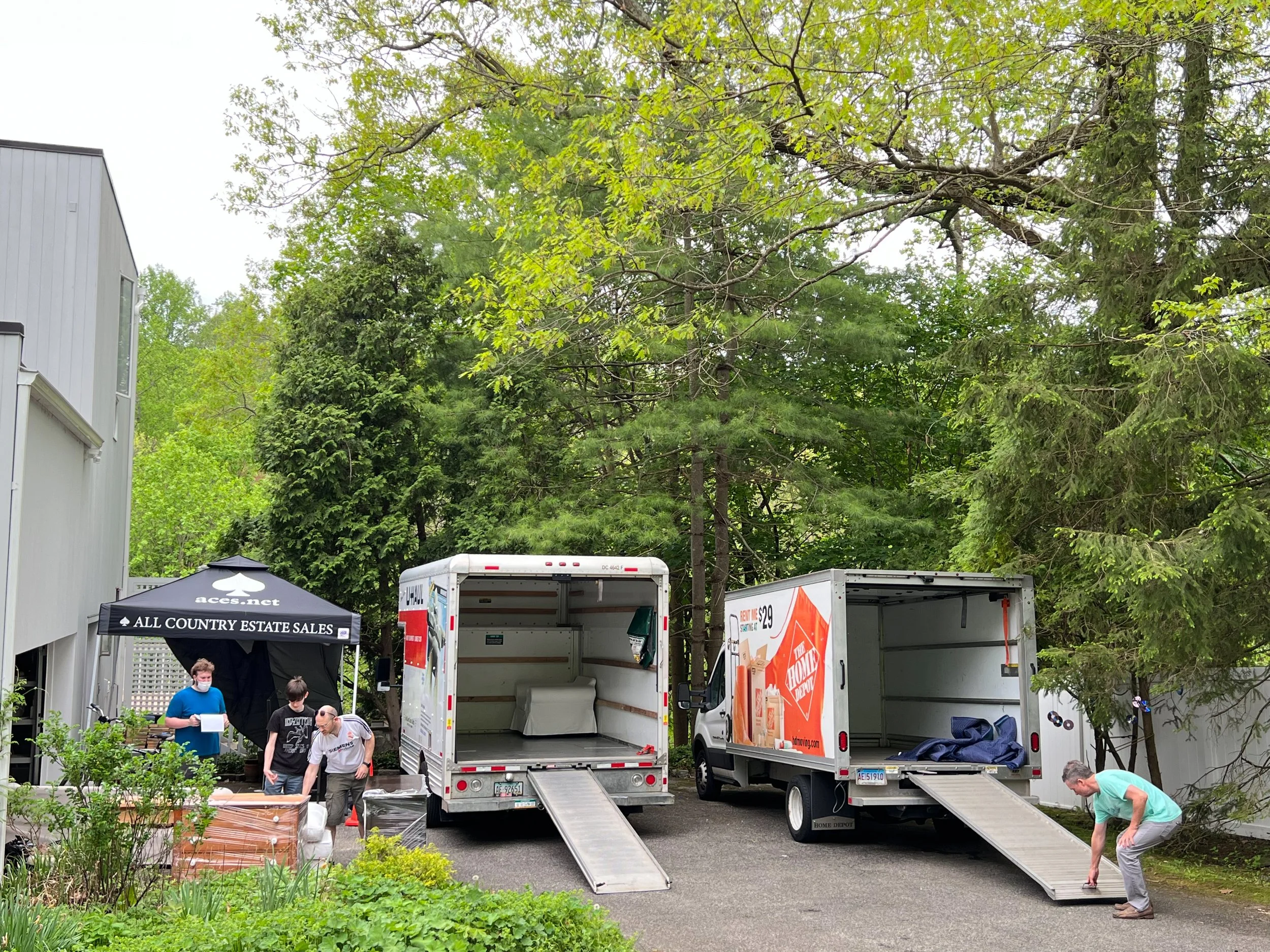 People loading moving trucks under a green tree with a canopy of leaves, next to a black tent with 'ALL COUNTRY ESTATE SALES' written on it, and a residential building, showing an ACES Online Estate Sale pickup day.