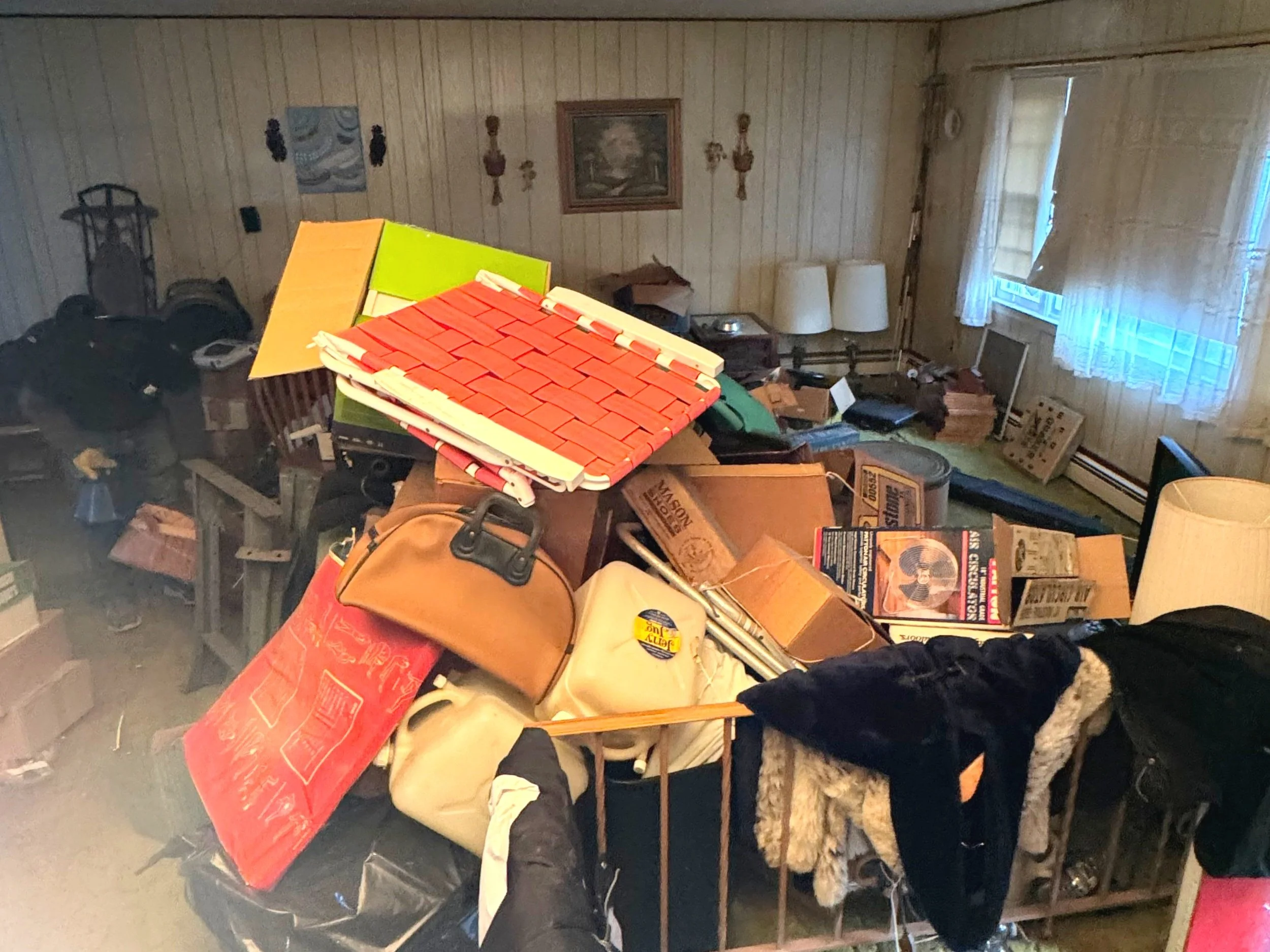 A cluttered room filled with various household items, including boxes, bags, furniture, and a lamp, with wooden walls and a window with curtains in the background, taken by ACES before starting an estate cleanout project.
