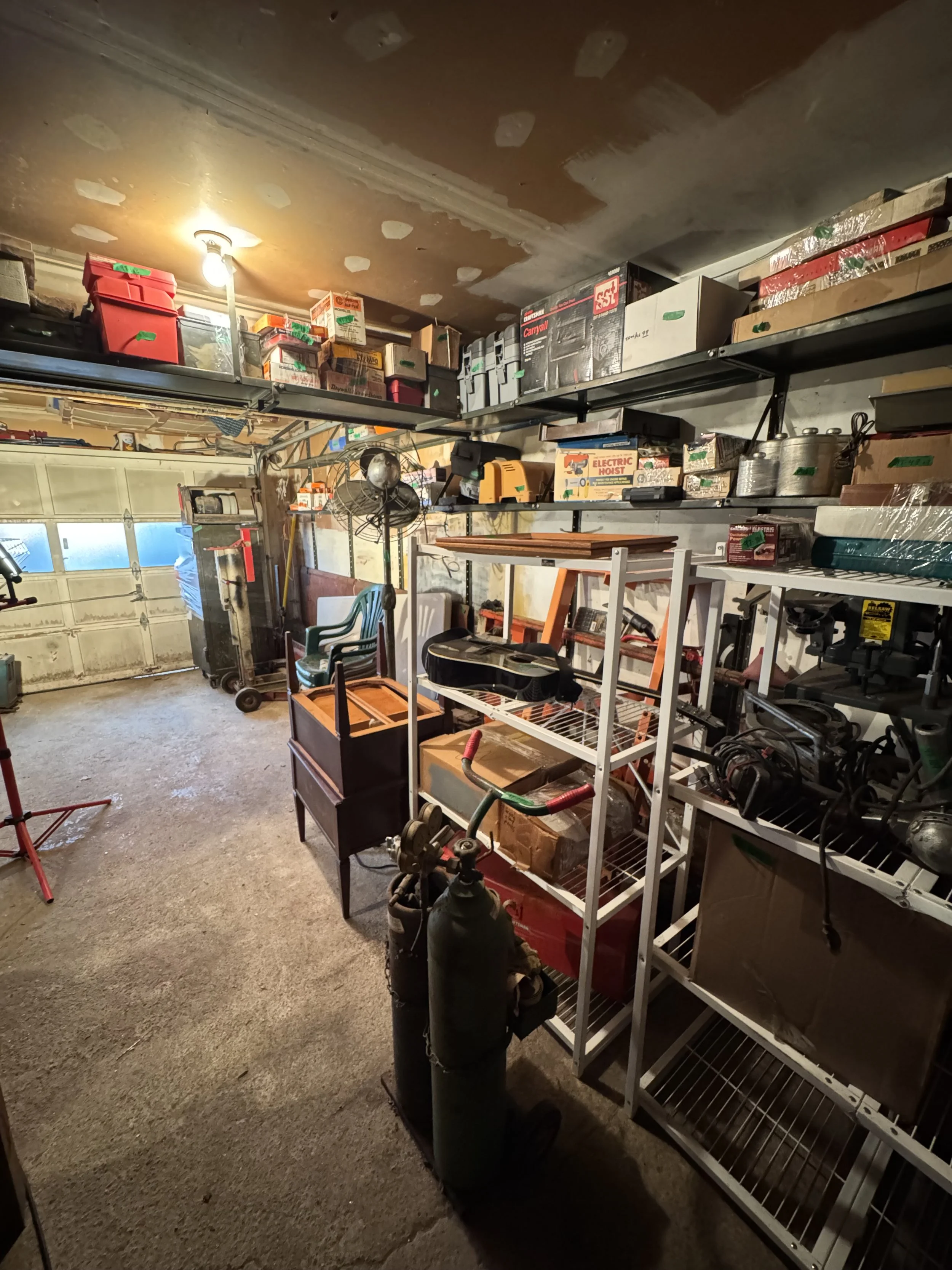 A full garage with storage shelves filled with boxes, tools, and miscellaneous items, a gas cylinder, plastic chairs, and a partially open garage door.