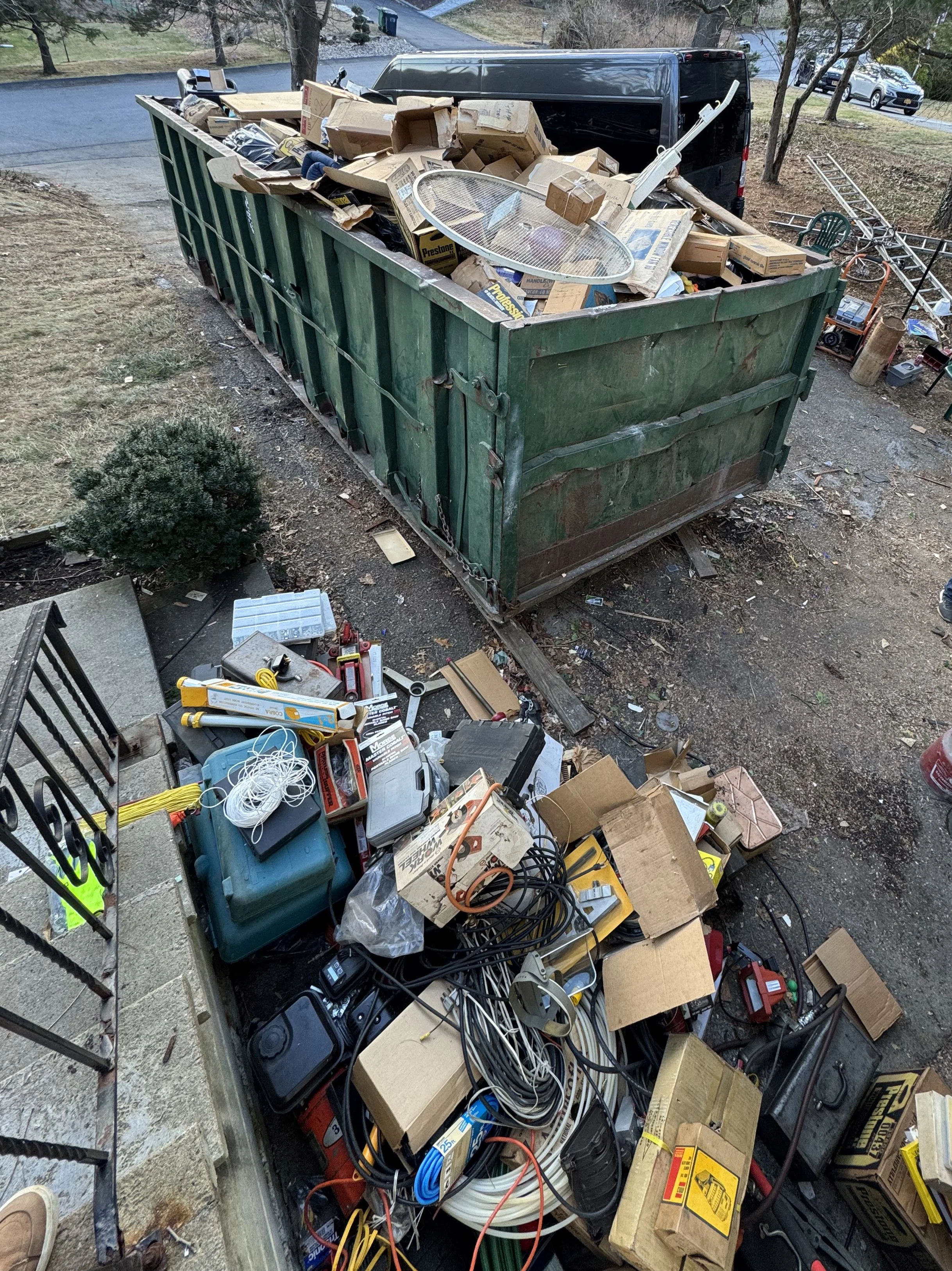 A large green dumpster filled with cardboard boxes and various discarded household items, with scattered tools and clutter on the ground nearby.
