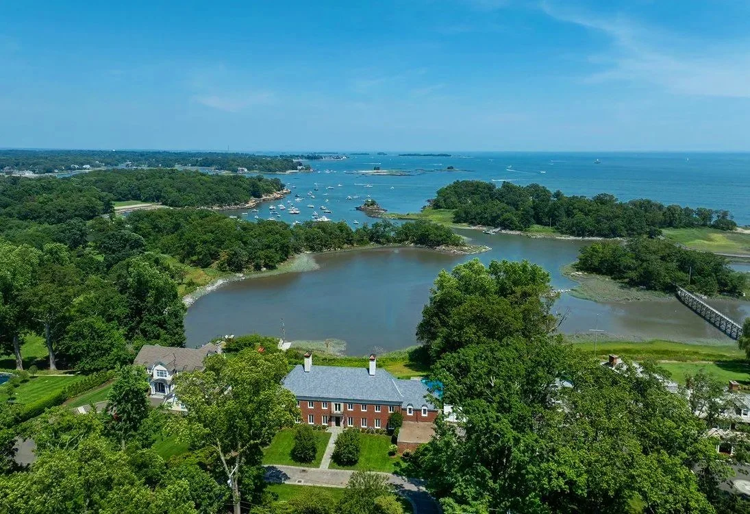 Aerial view of a coastal landscape featuring a lake surrounded by lush green trees, houses, and a bridge, with the expansive ocean in the background and numerous boats on the water, showing an ACES estate client's home.