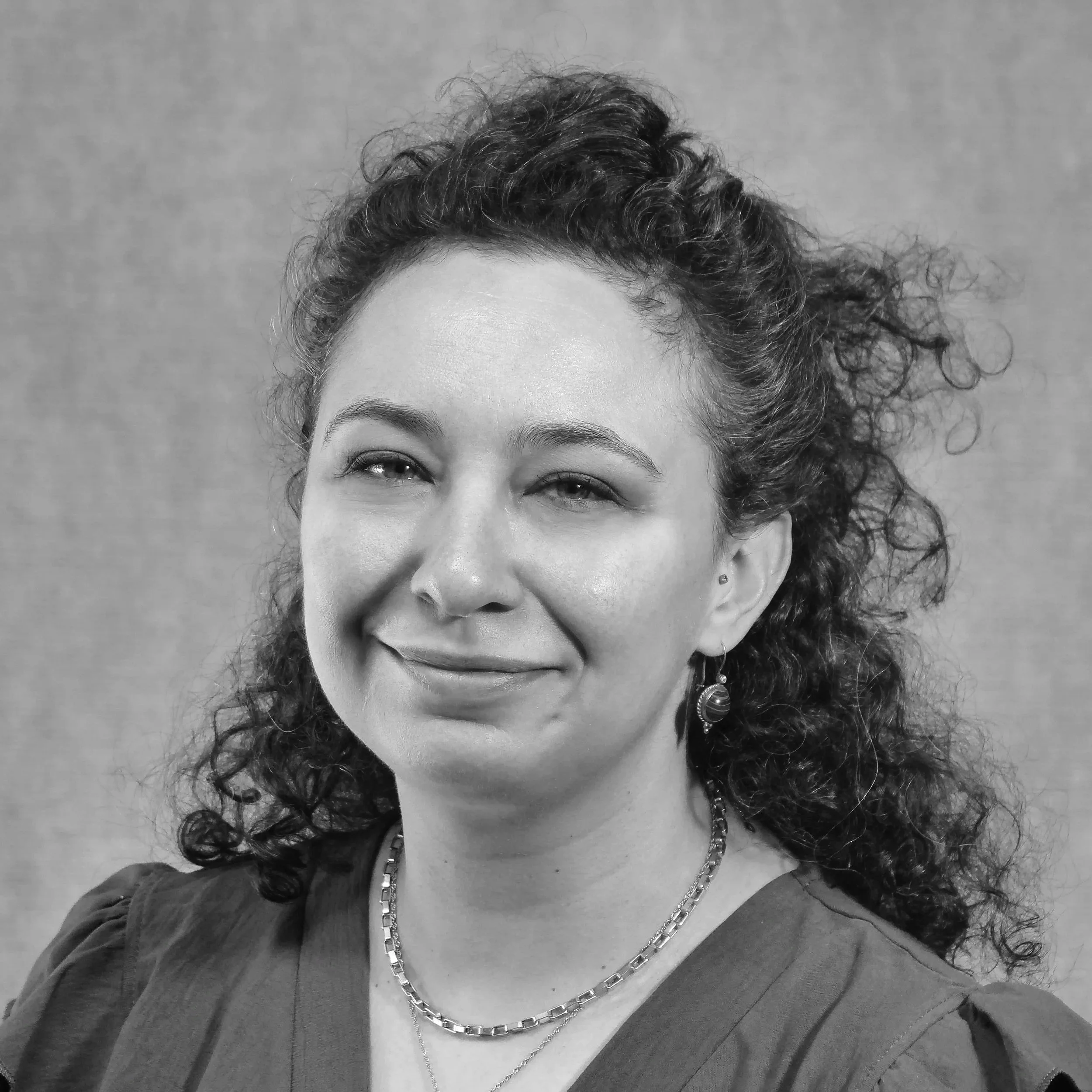 A woman with shoulder-length black hair smiling in front of green plants.