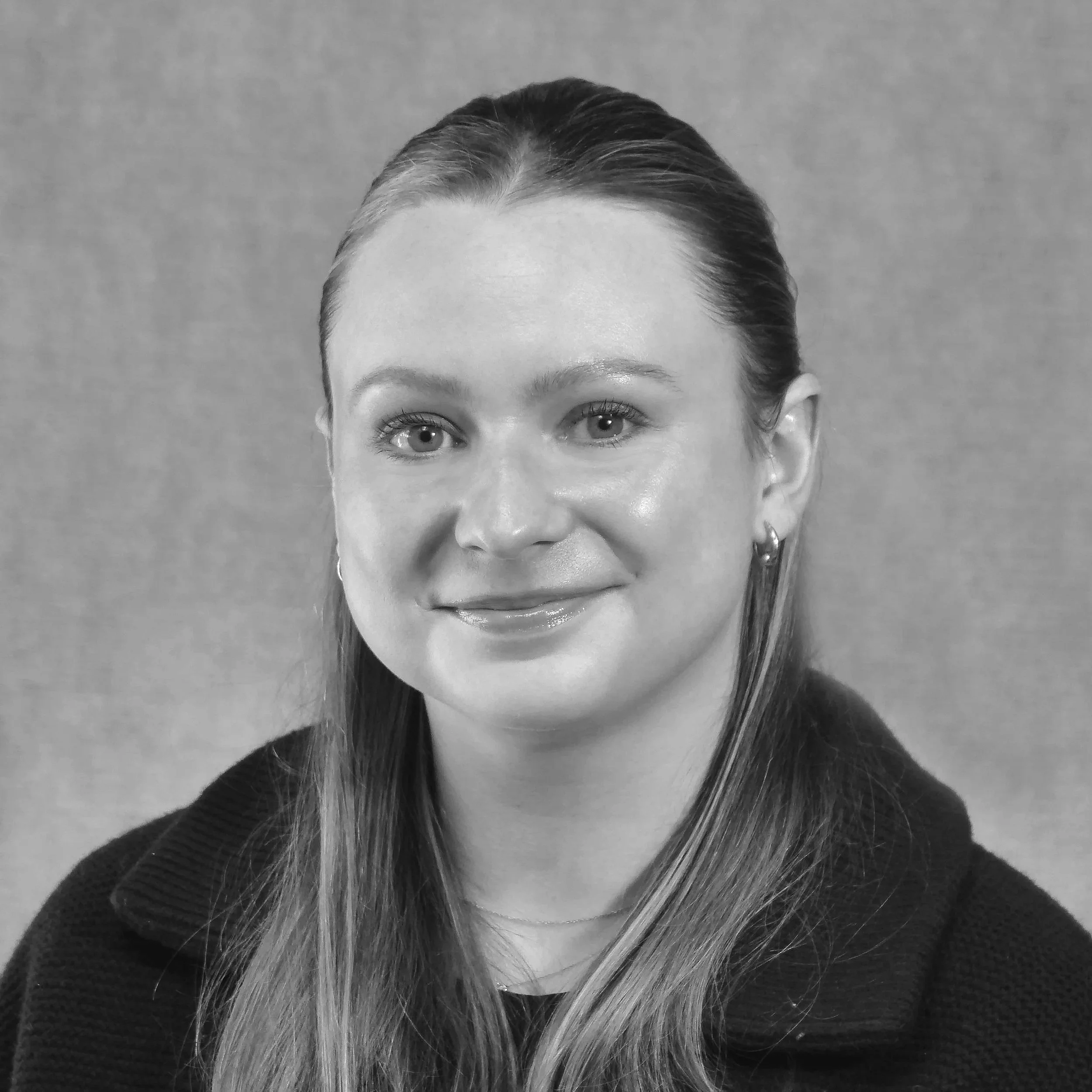 A smiling young woman with black hair wearing a blue top, standing among green plants in a greenhouse.