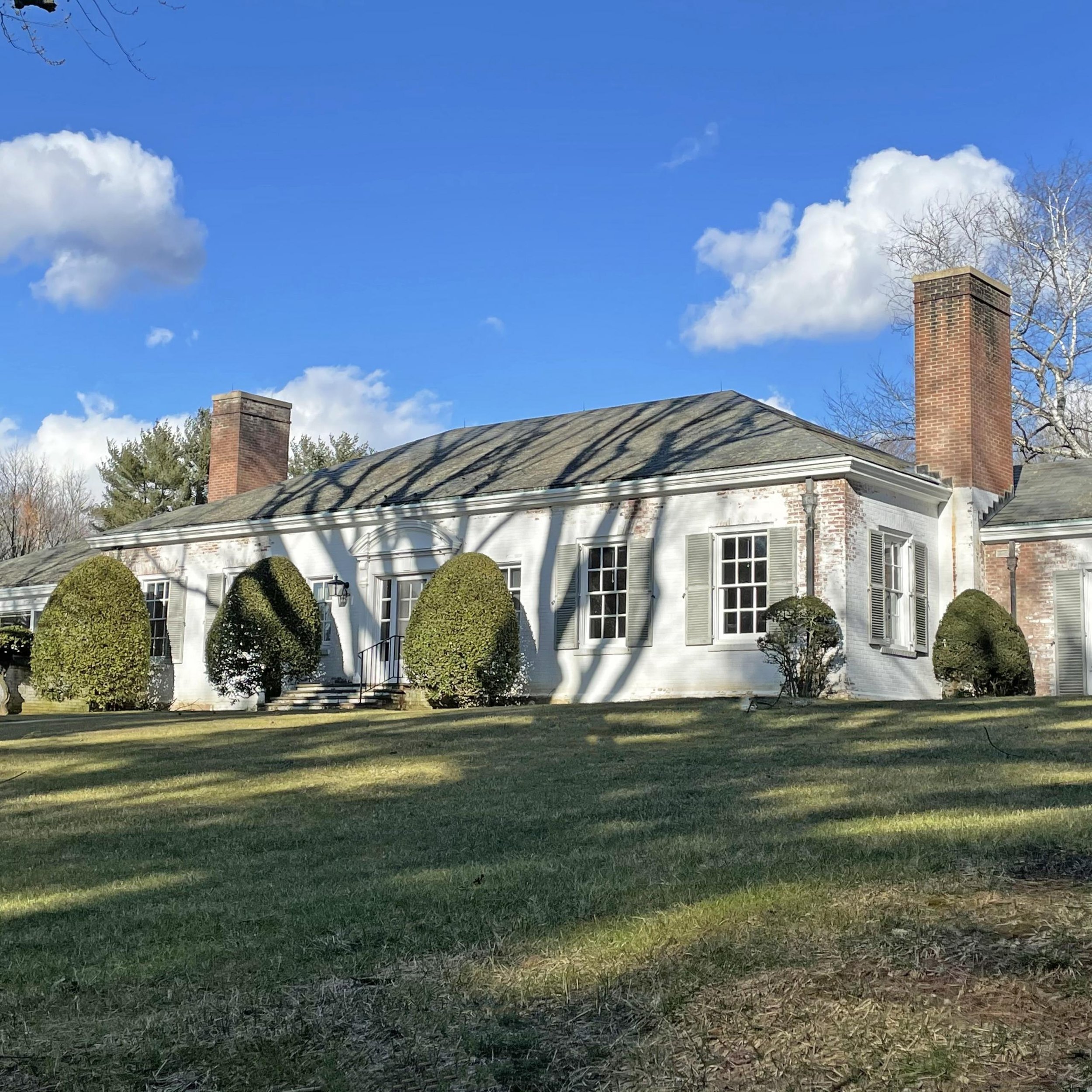 White house with multiple windows, surrounded by bushes, under a blue sky with clouds, with shadows cast on the house, representing an ACES estate client's home.