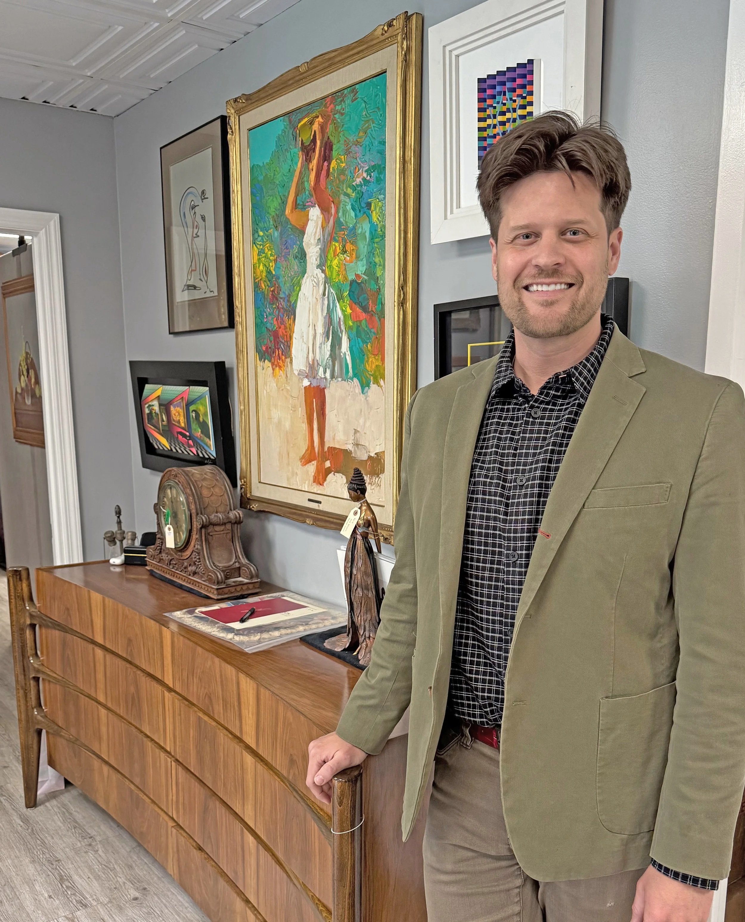 A man with short brown hair and a beard smiling, dressed in a green blazer and a black checkered shirt, standing in an art gallery or home, beside a wooden sideboard decorated with antique clocks, sculptures, and artwork on the wall behind him.