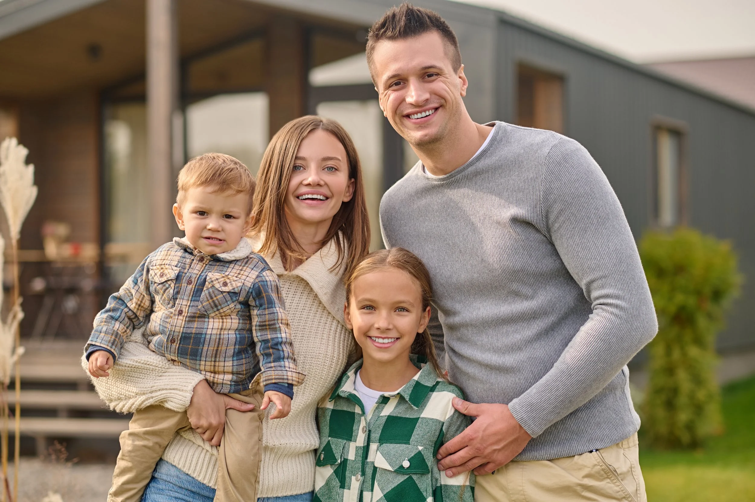 A smiling family of four standing outside in front of a modern house with a porch. They include a man, a woman, and two children, a boy and a girl, all dressed in casual fall clothing.
