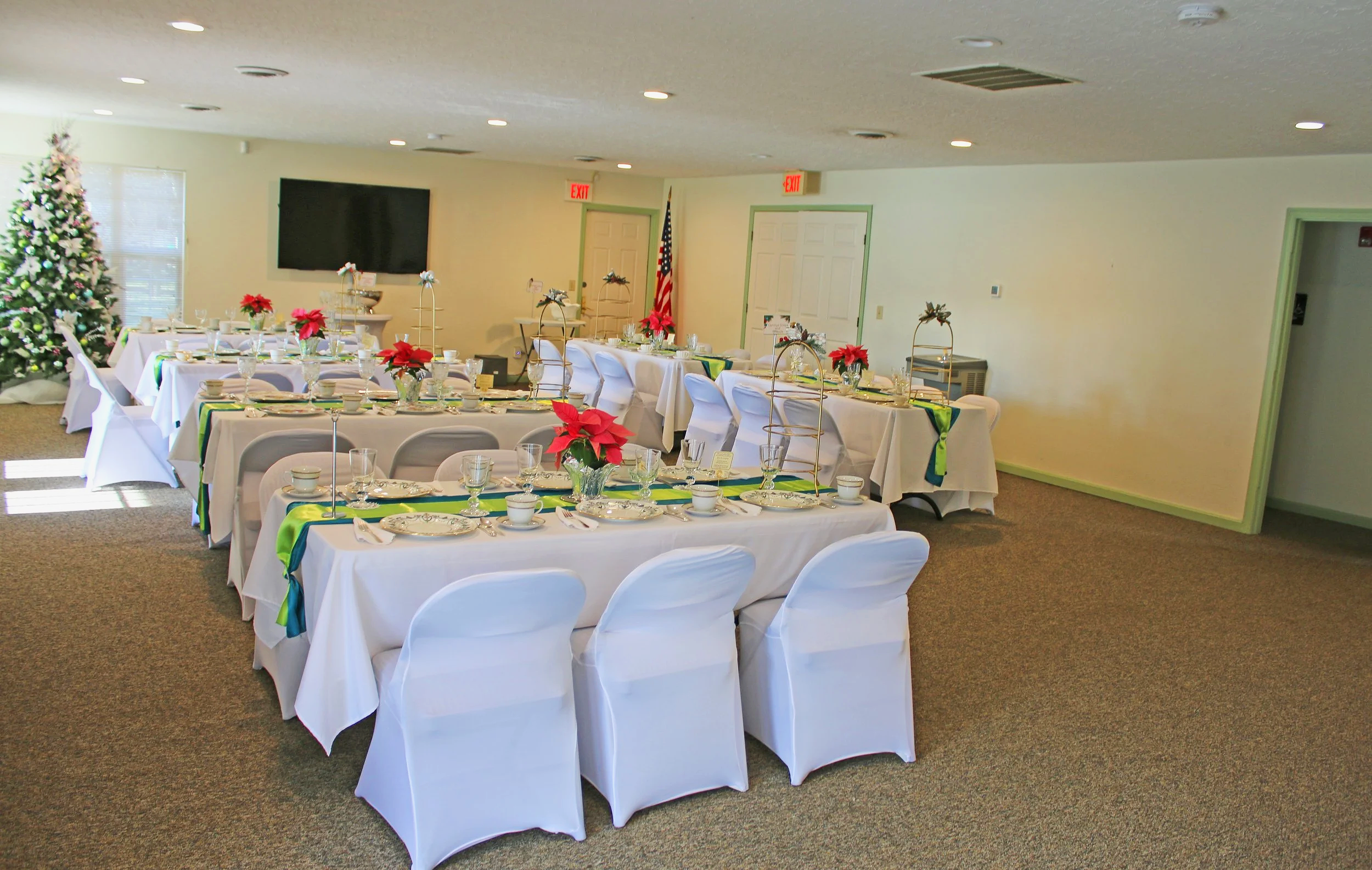 A decorated banquet hall with Christmas trees, round and rectangular tables covered with white tablecloths, poinsettia floral arrangements, and white chairs, set for a holiday celebration.