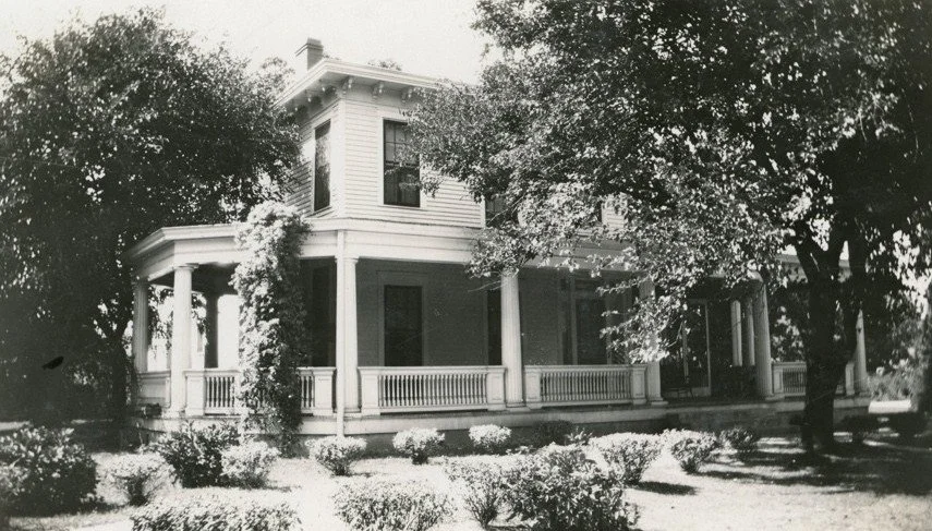 A large historic house with a wraparound porch, multiple columns, and Victorian architectural details, surrounded by trees and shrubbery.