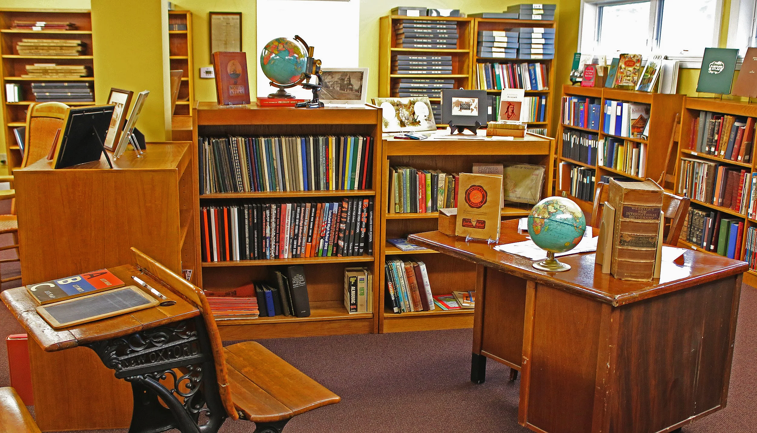 Interior of a bookstore or library with wooden shelves filled with books, globes, framed pictures, and reading materials on tables, with chairs and windows in the background.