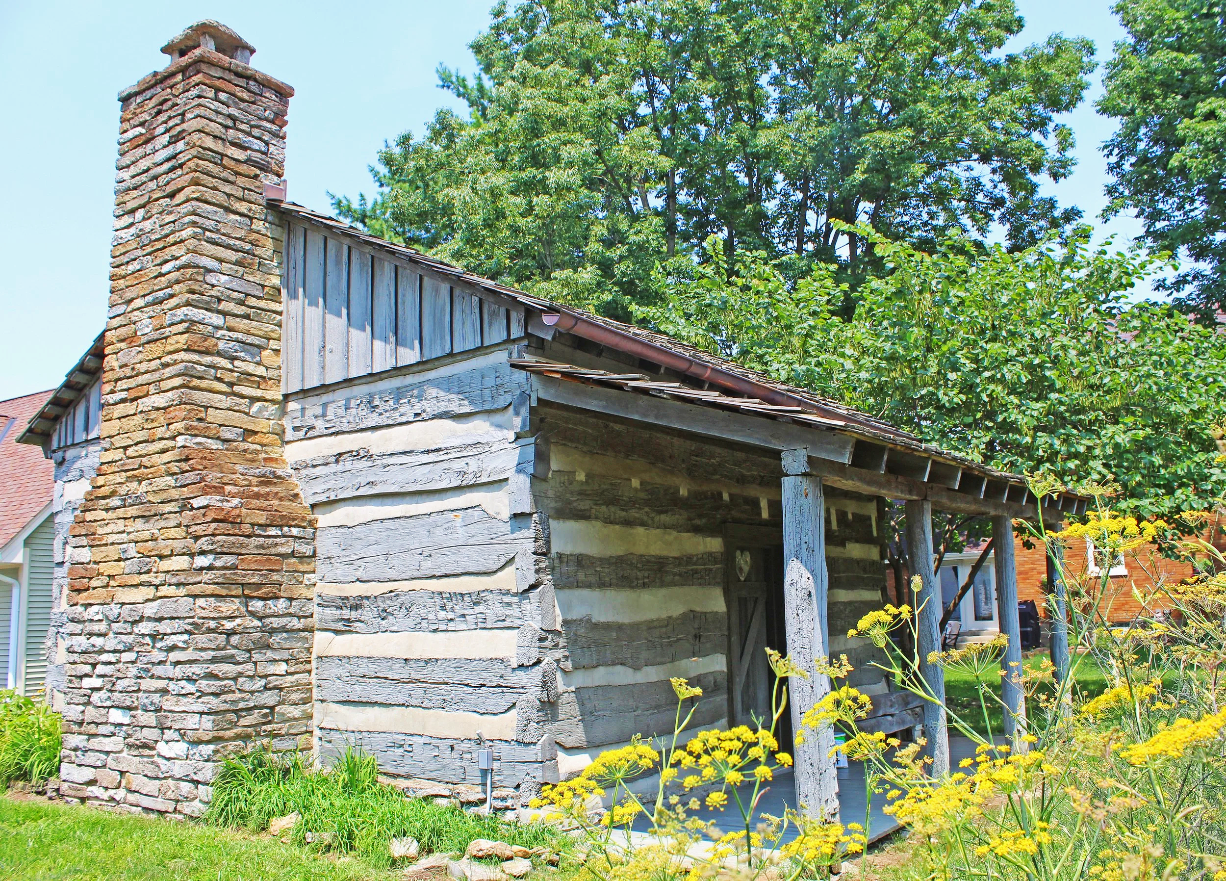 An old wooden shed with a brick chimney in a yard surrounded by green trees and yellow flowering plants.