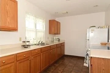 An empty kitchen with wooden cabinets, a white countertop, a white refrigerator, and a window with sheer curtains. Contains a sink, stove with oven, and microwave.