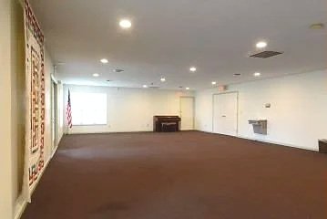 Empty room with white walls, brown carpet, and ceiling lights, featuring a window and a decorative wall hanging. Also has a screen to show videos and images.