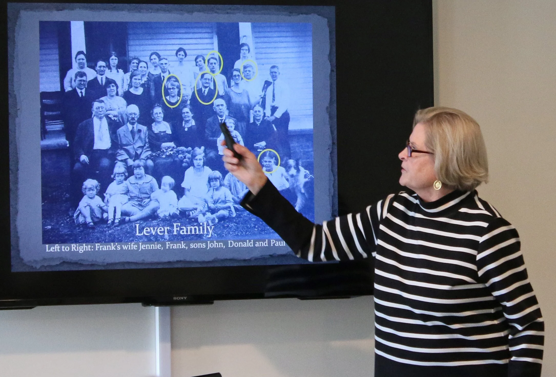 Woman pointing at a family photo on a screen during a presentation. The photo shows a group of people with some of them circled in yellow. The text below the photo reads 'Lever Family' and names of individuals, including Jennie, Frank, John, Donald, and Paul.