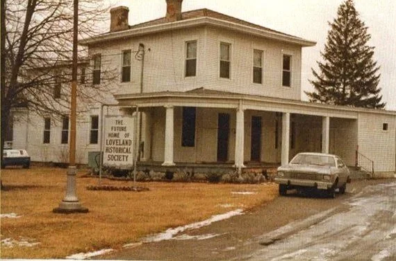 Historic two-story white house with a porch, sign indicating it's the future home of Loveland Historical Society, parked car and muddy driveway in front, on a cloudy day.