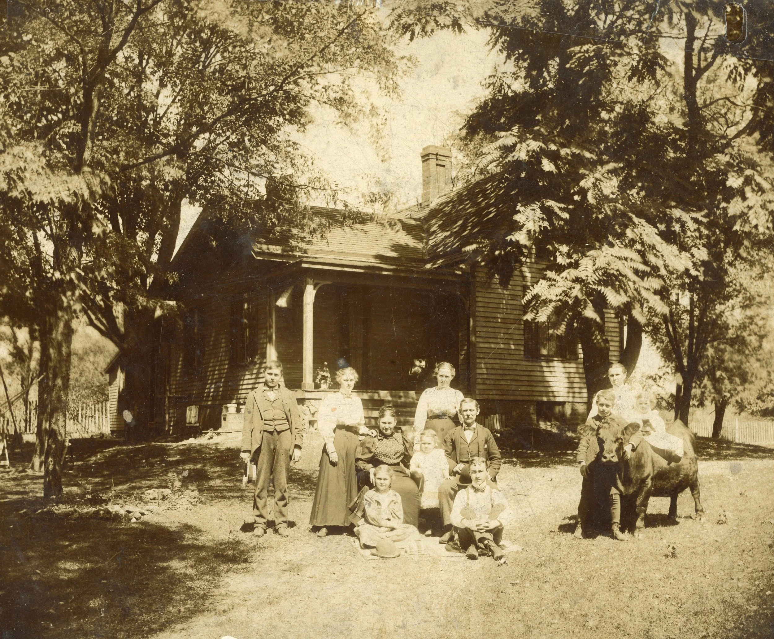 A sepia-toned photograph of ten people, including children and adults, gathered in front of a wooden house with trees surrounding it. Some are sitting, some standing, with one person holding a small animal, possibly a dog or a pig. The scene appears to be from the late 19th or early 20th century.