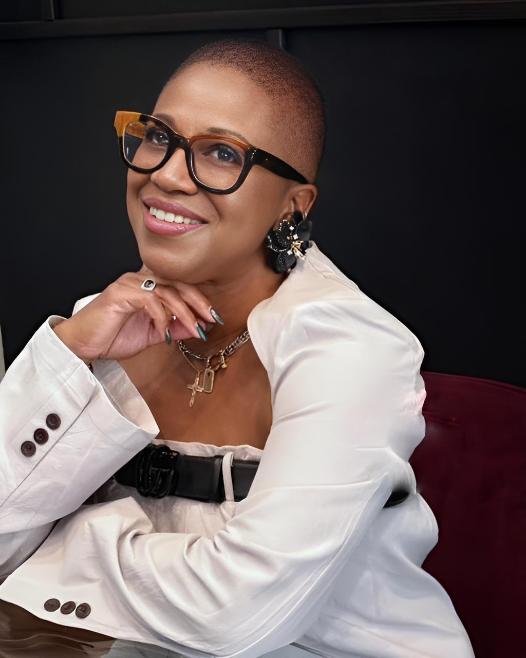 A woman with short hair, glasses, and earrings, dressed in a white shirt, smiling and resting her chin on her hand.