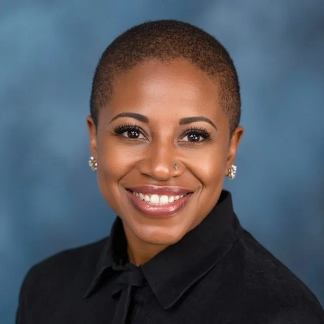 Close-up portrait of a smiling Black woman with a short buzz cut hairstyle, wearing a black collared shirt and pearl earrings, against a blue background.