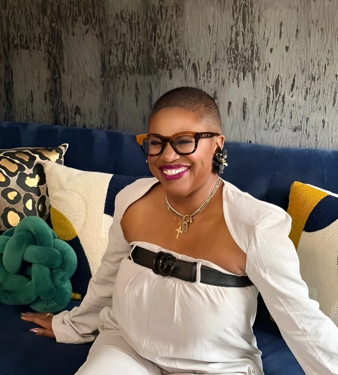 A smiling woman with short hair, wearing glasses, gold jewelry, and a white off-shoulder top with a black belt, sitting on a blue couch with patterned pillows and a decorative knot pillow.