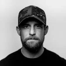 A man with a beard wearing a camouflage baseball cap and a black shirt, looking at the camera in a black-and-white portrait.