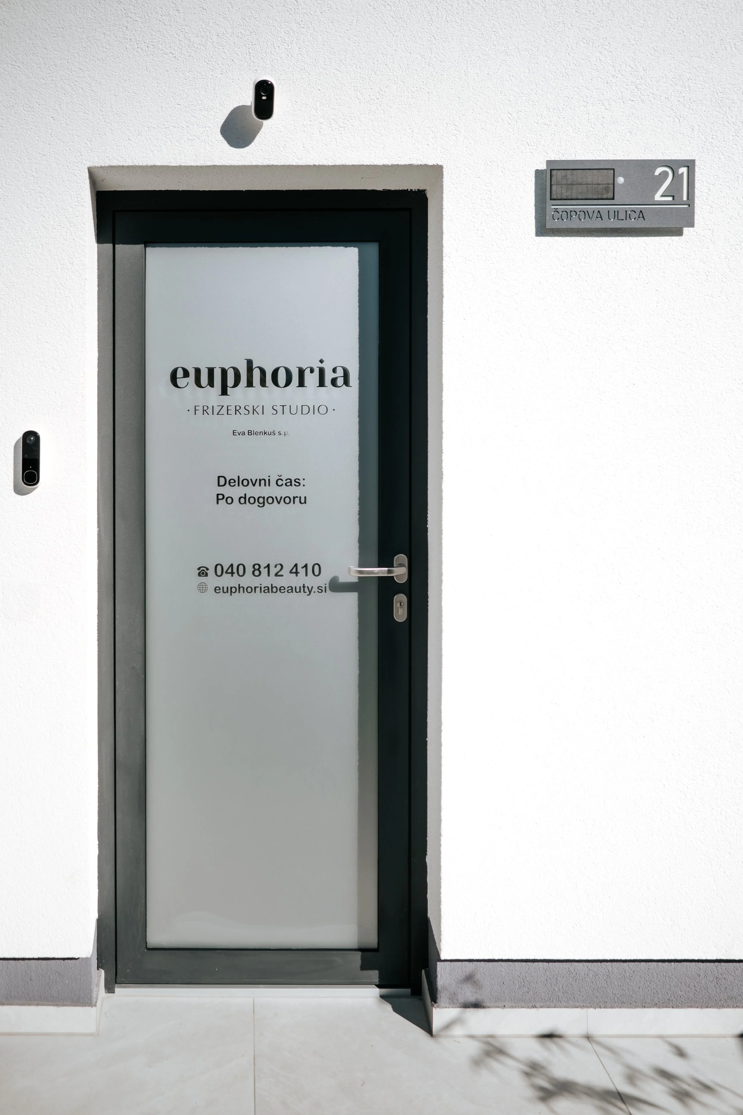 White door with frosted glass panel advertising euphoria beauty salon, with contact information and hours of operation, set in a white building wall. To the right, a street number and sign indicating Cnopova Ulica.