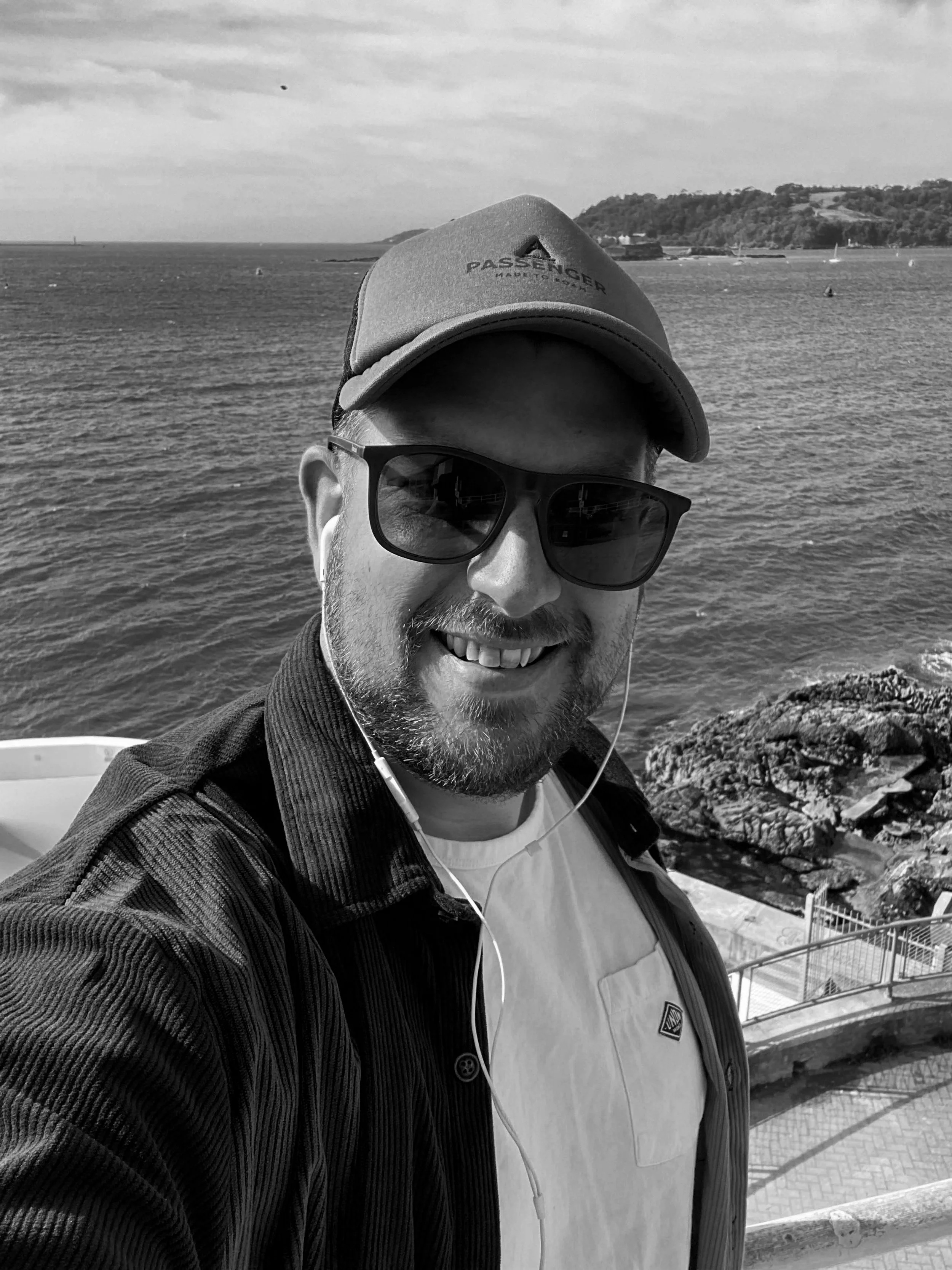 A man taking a selfie near the water with a rocky shoreline and distant land in the background.