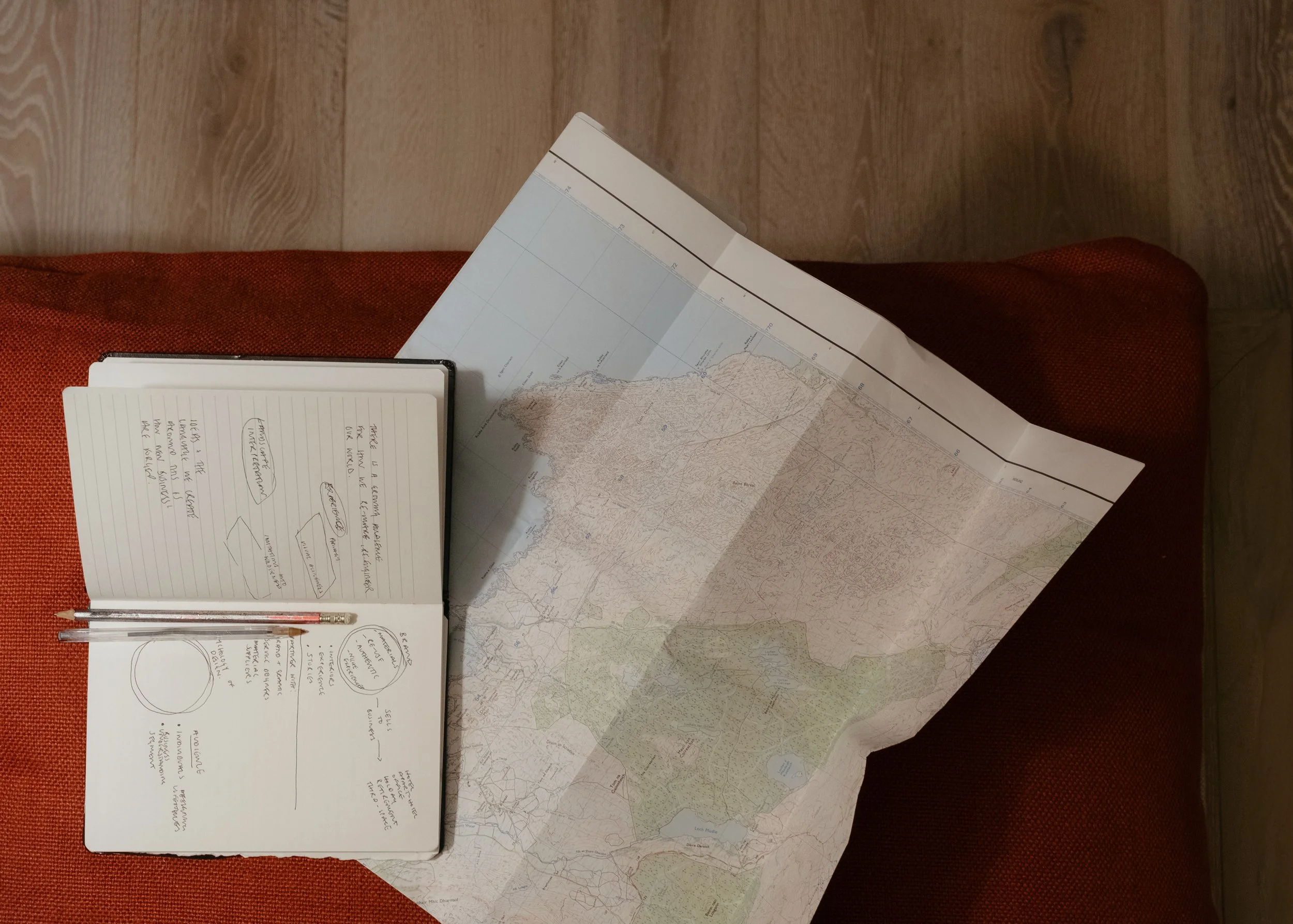 Open notebook with handwritten notes and pen, and large folded map on a red textured chair.