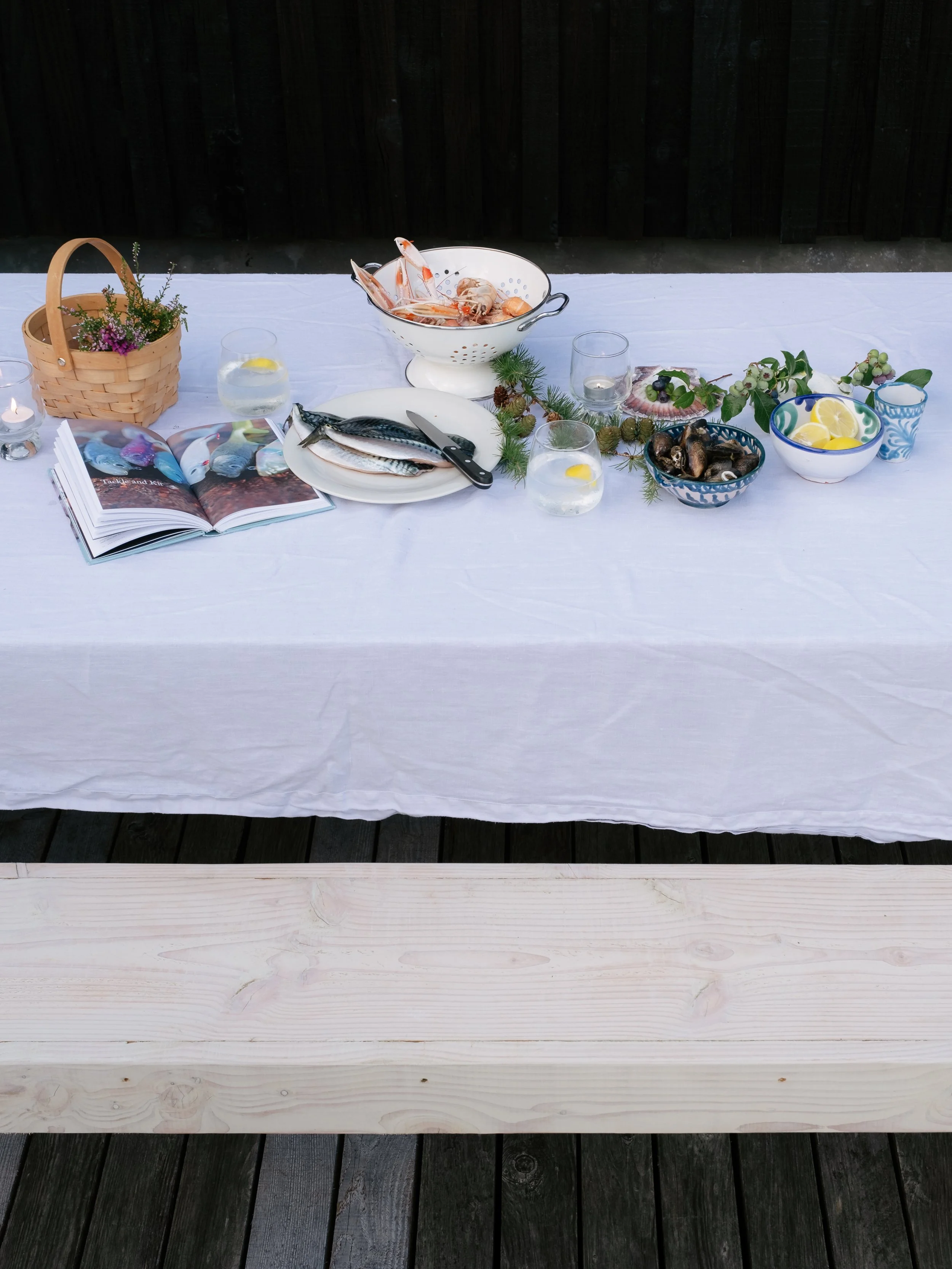 Table outdoors with seafood, a cookbook, candles, a basket of flowers, and decorative greenery on a white tablecloth, with a dark wooden fence in the background.