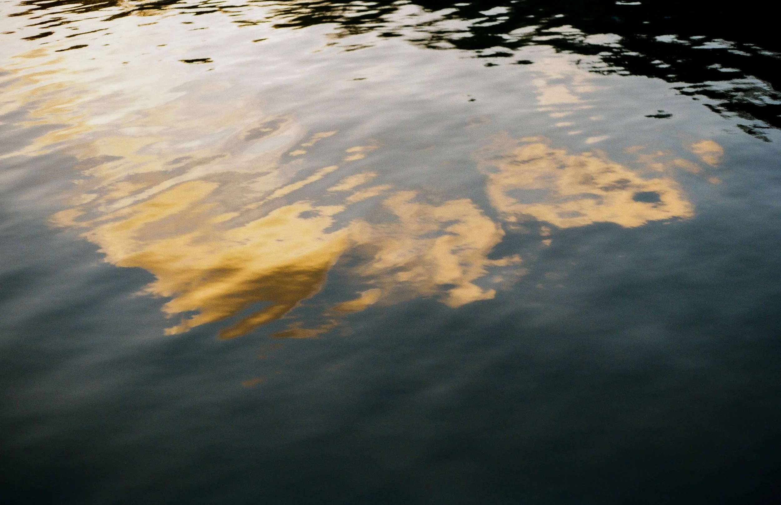 Reflection of a cloudy sky with sunlight on the surface of a dark body of water.
