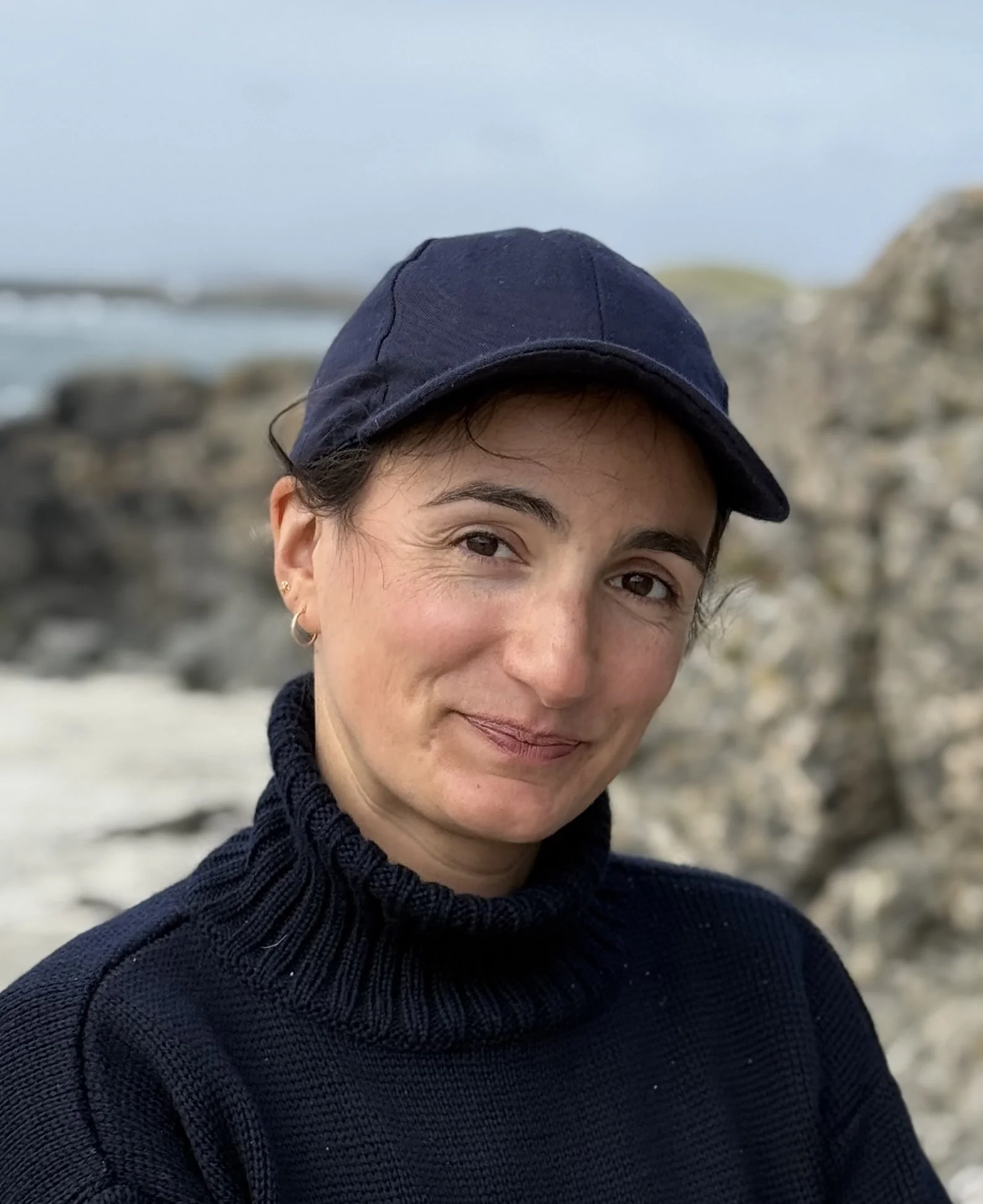Close-up of a woman wearing a navy blue cap and black sweater, outdoors near rocks with blurred background.