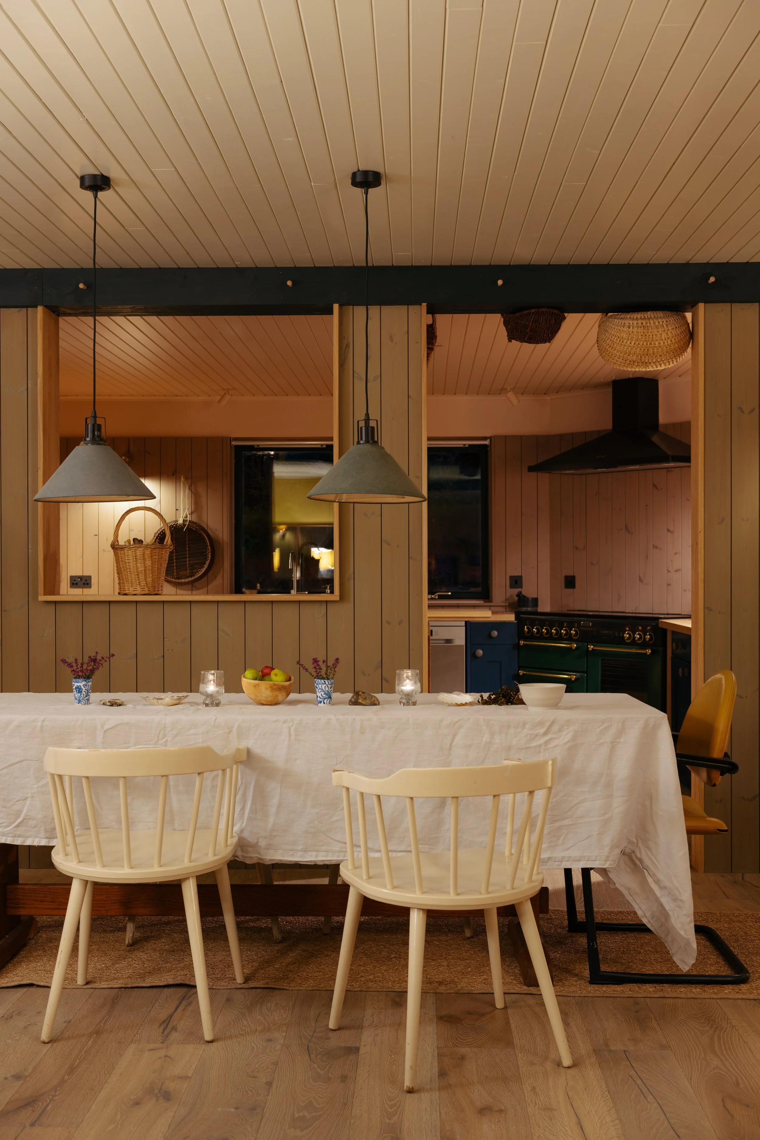 A dining table set for a meal with a white tablecloth, bowls of fruit and flowers in small vases, and candles. Wooden chairs surround the table. The background features a wooden-paneled kitchen with hanging ceiling lights, a basket on a shelf, a window, and visible kitchen appliances.