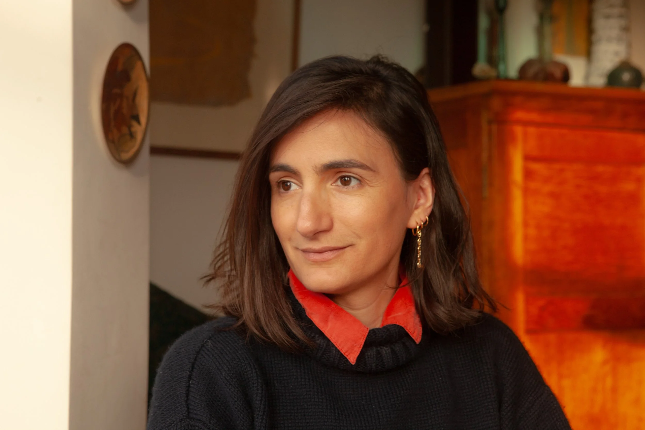 A woman with dark brown hair, wearing a black sweater with a red collar, standing indoors near a white wall and a wooden cabinet with decorative items.