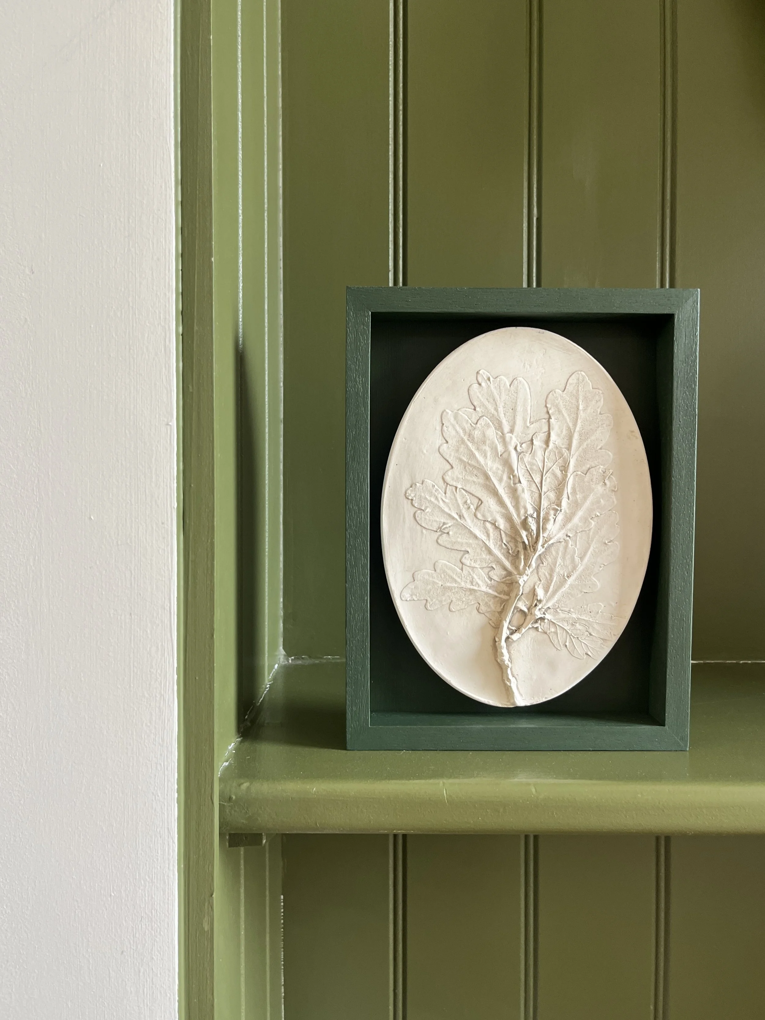 A framed white ceramic plate with a relief carving of a tree branch and leaves, displayed in a green wall-mounted frame.