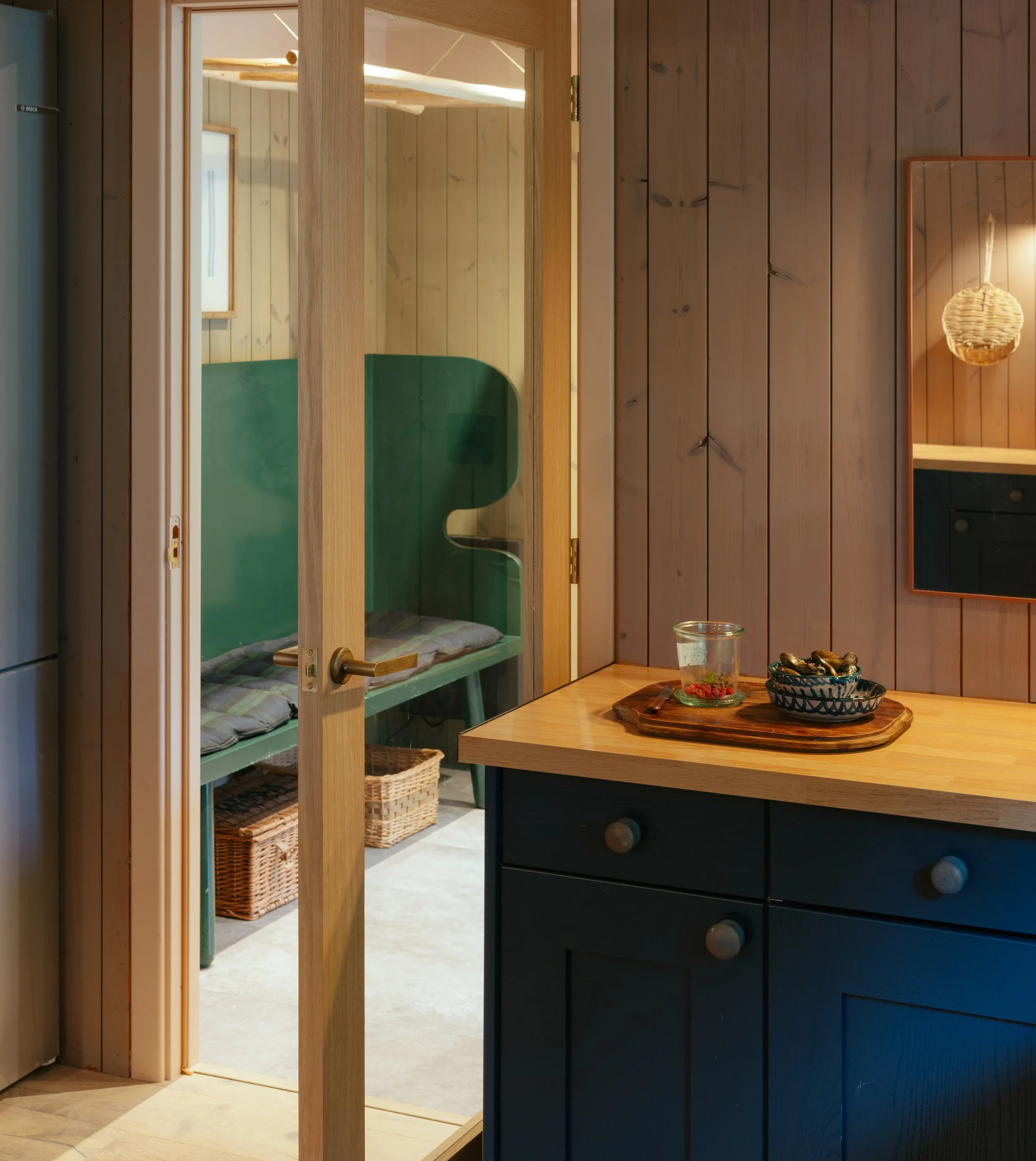 A view through an open door into a cozy room with light wooden walls, featuring a green seating area with cushions, and a basket on the floor. Part of the blue kitchen cabinet with round wooden knobs is visible in the foreground.
