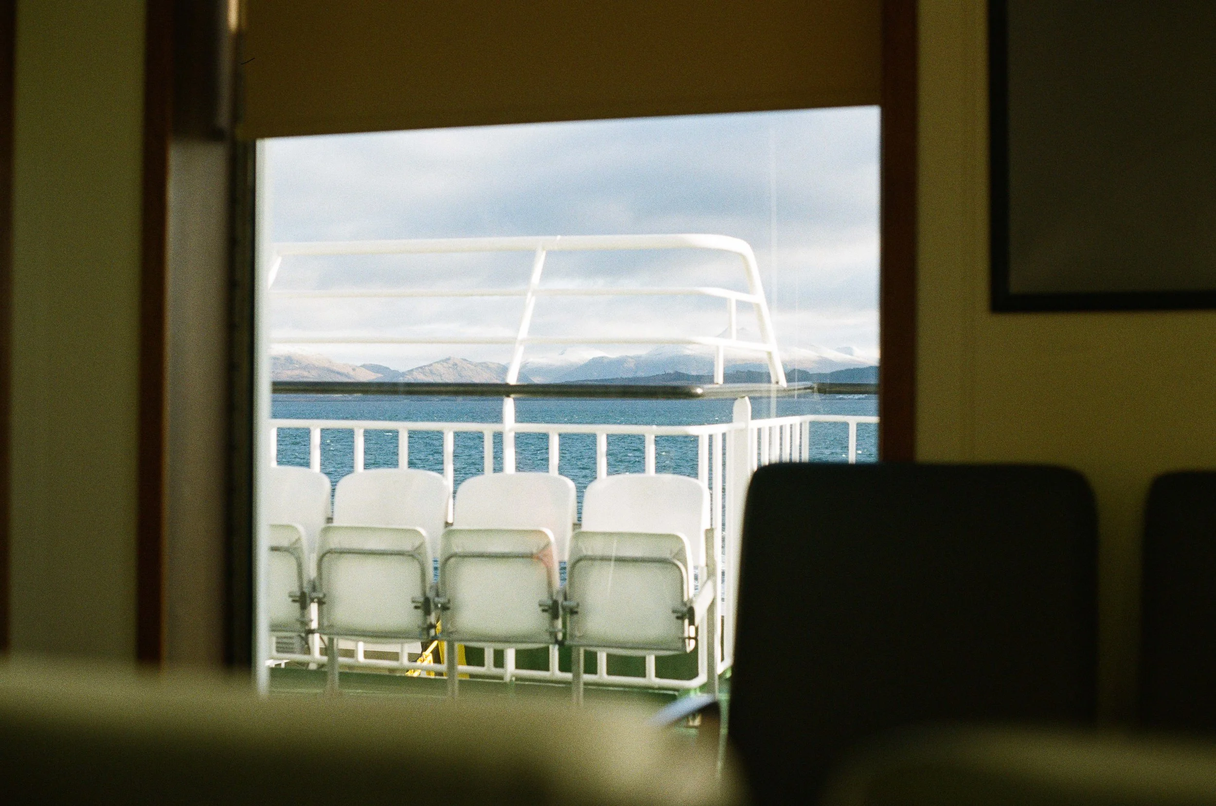 A room viewed through a door or window with a blurred foreground. Outside, there are white chairs placed on a deck overlooking a body of water with mountains in the distance under a cloudy sky.