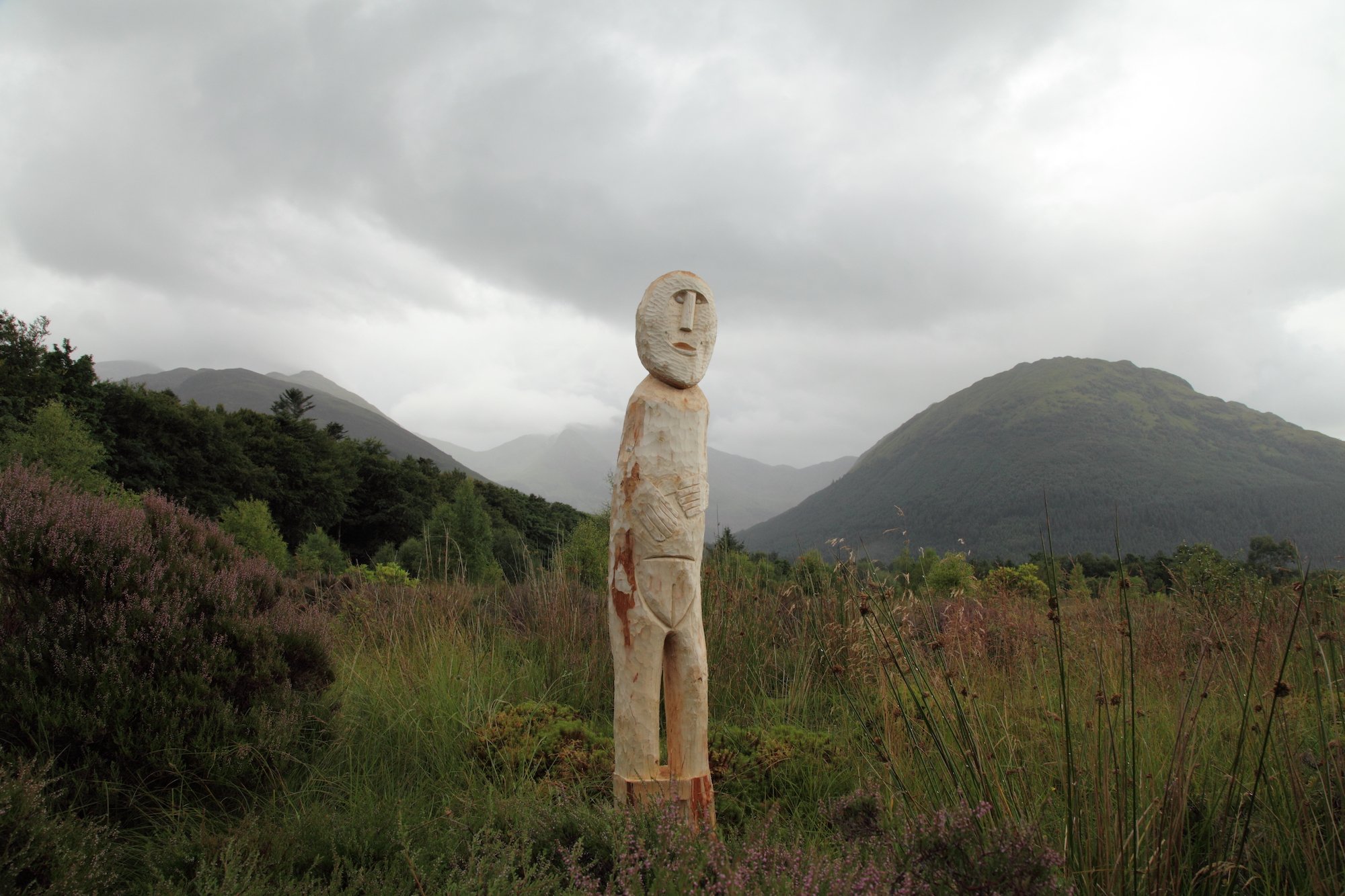 Wooden sculpture of a human figure with an elongated face stands in grassy field with mountains and cloudy sky in background.