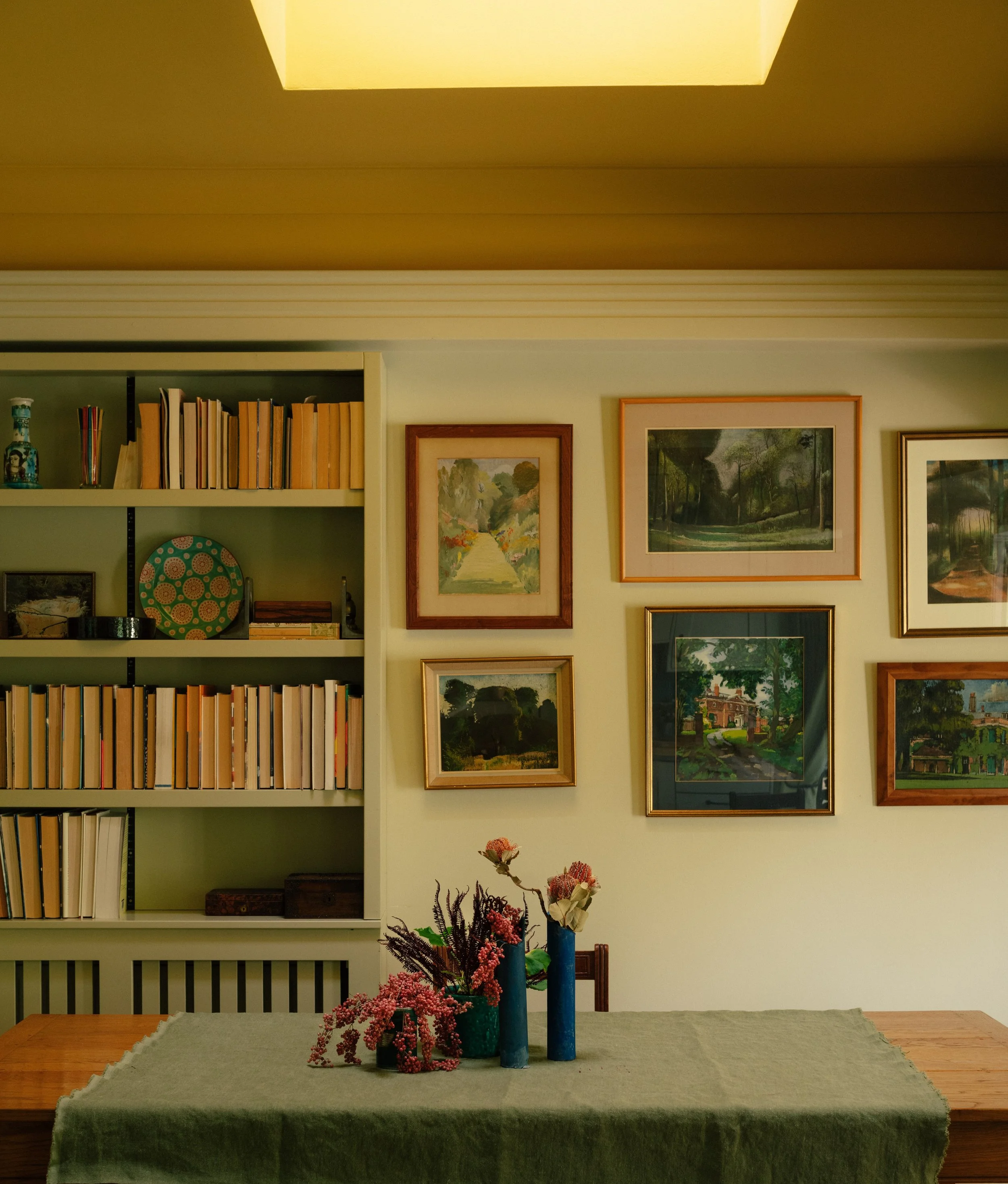 Living room with a shelf filled with books and framed paintings on the wall, has a dining table with vases and flowers, and a ceiling skylight.