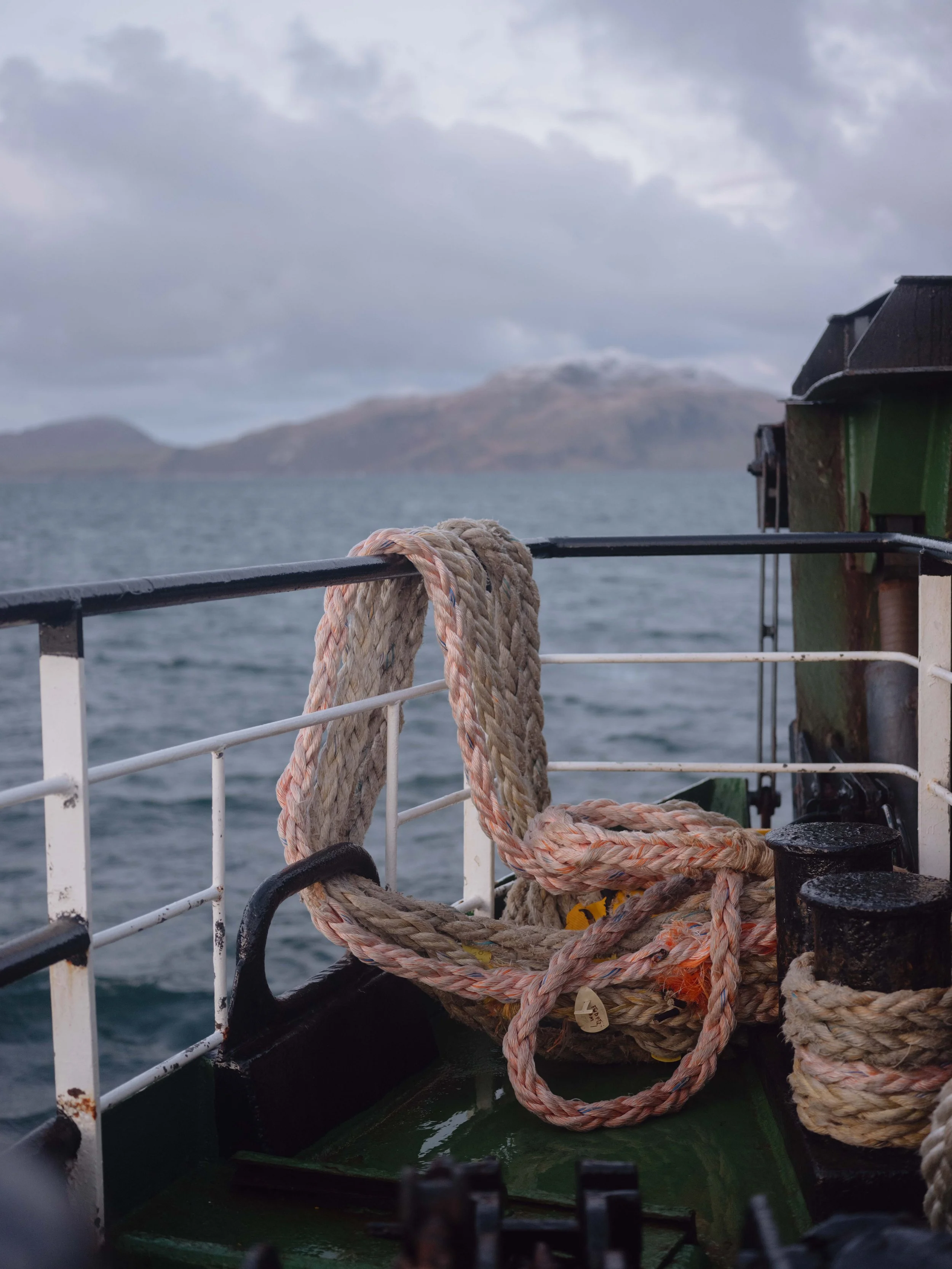 Close-up of a ship's deck with thick, knotted rope tied to the railing, overlooking a body of water with distant landforms and clouds in the sky.