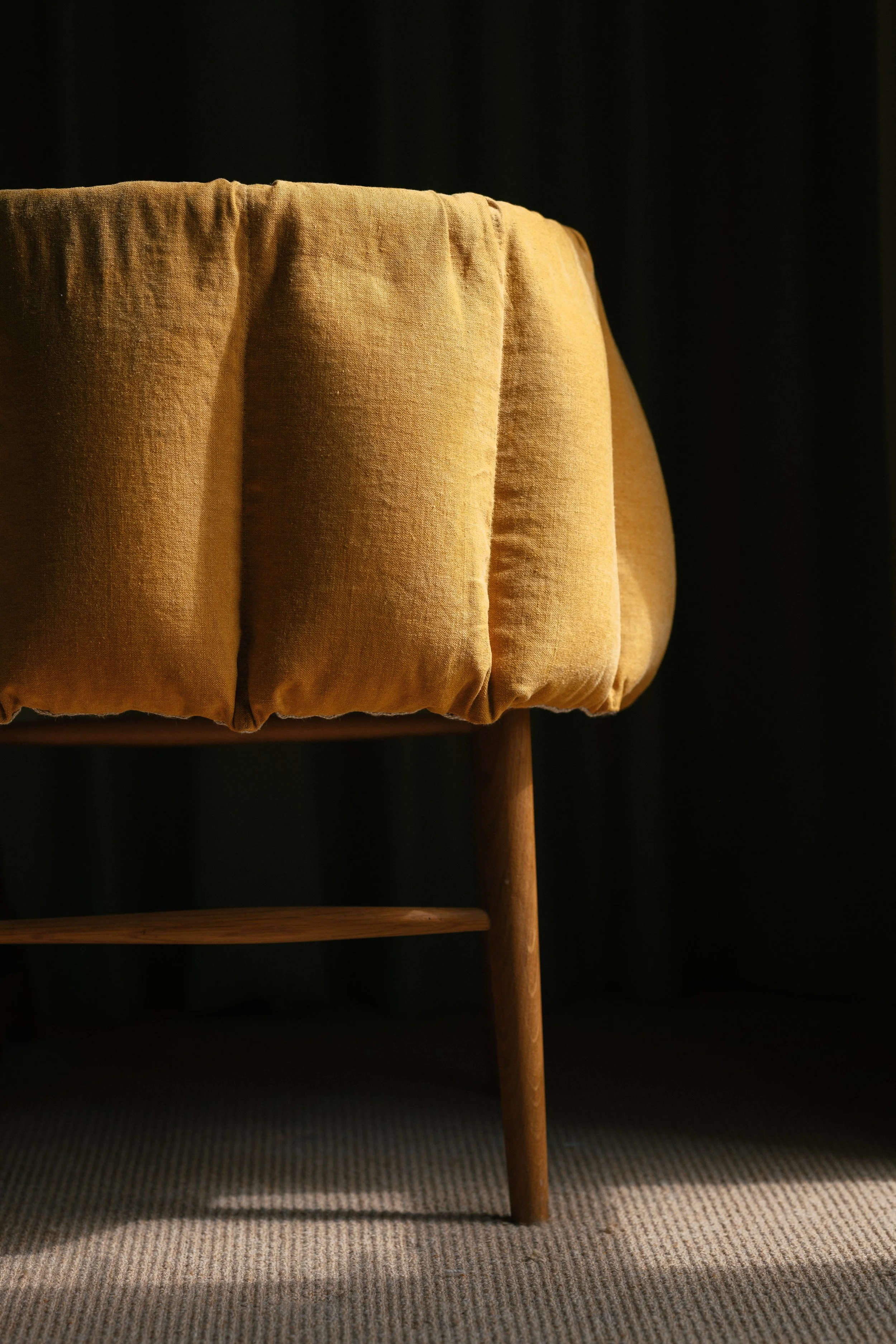 Close-up of a mustard-colored upholstered chair or bench with wooden legs, on a striped carpet, against a dark background.