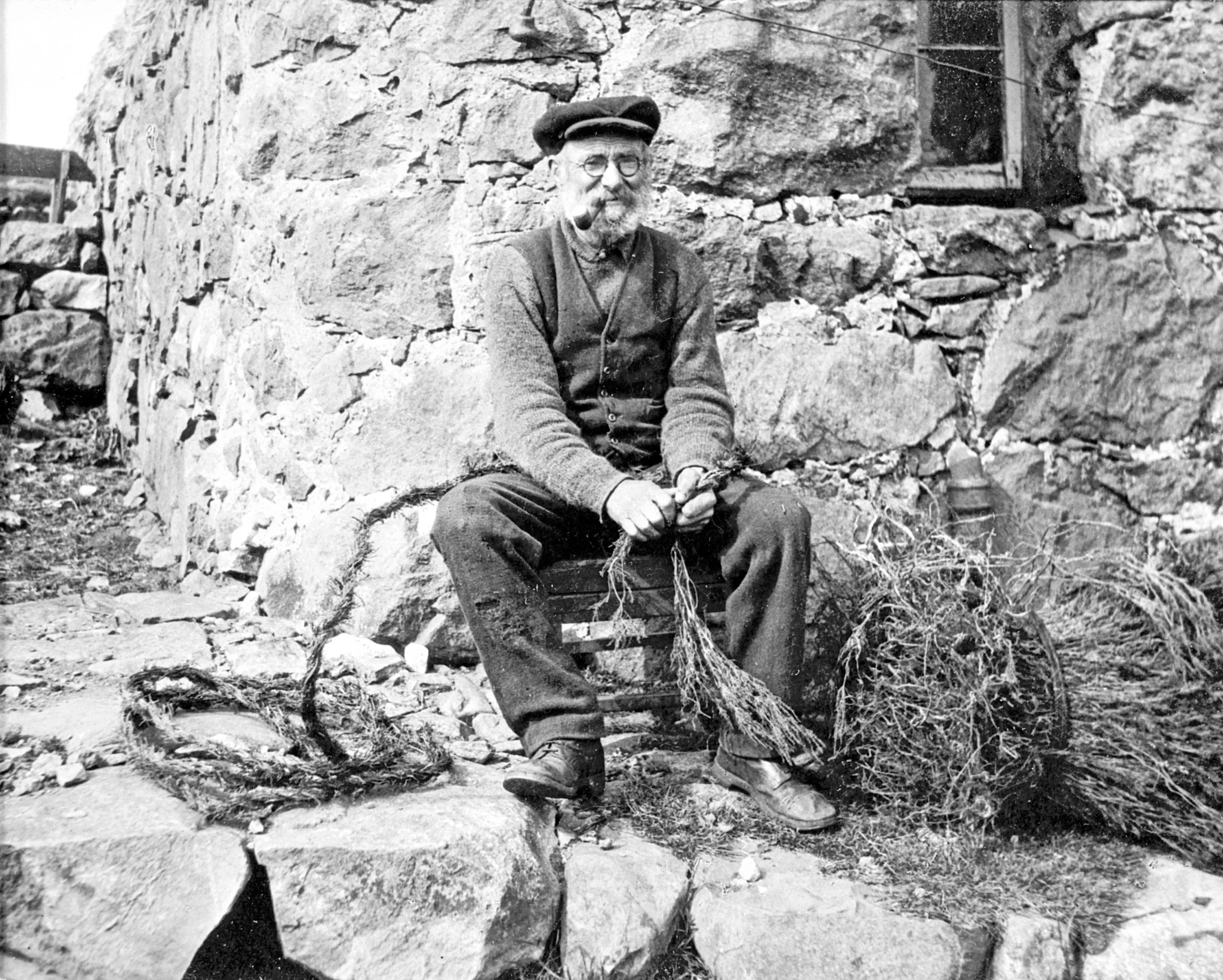 A black and white photo of an elderly man sitting on a small wooden stool against a stone wall. The man is holding some dried plants or weeds, is wearing glasses, a cap, and a suit with a pipe in his mouth.