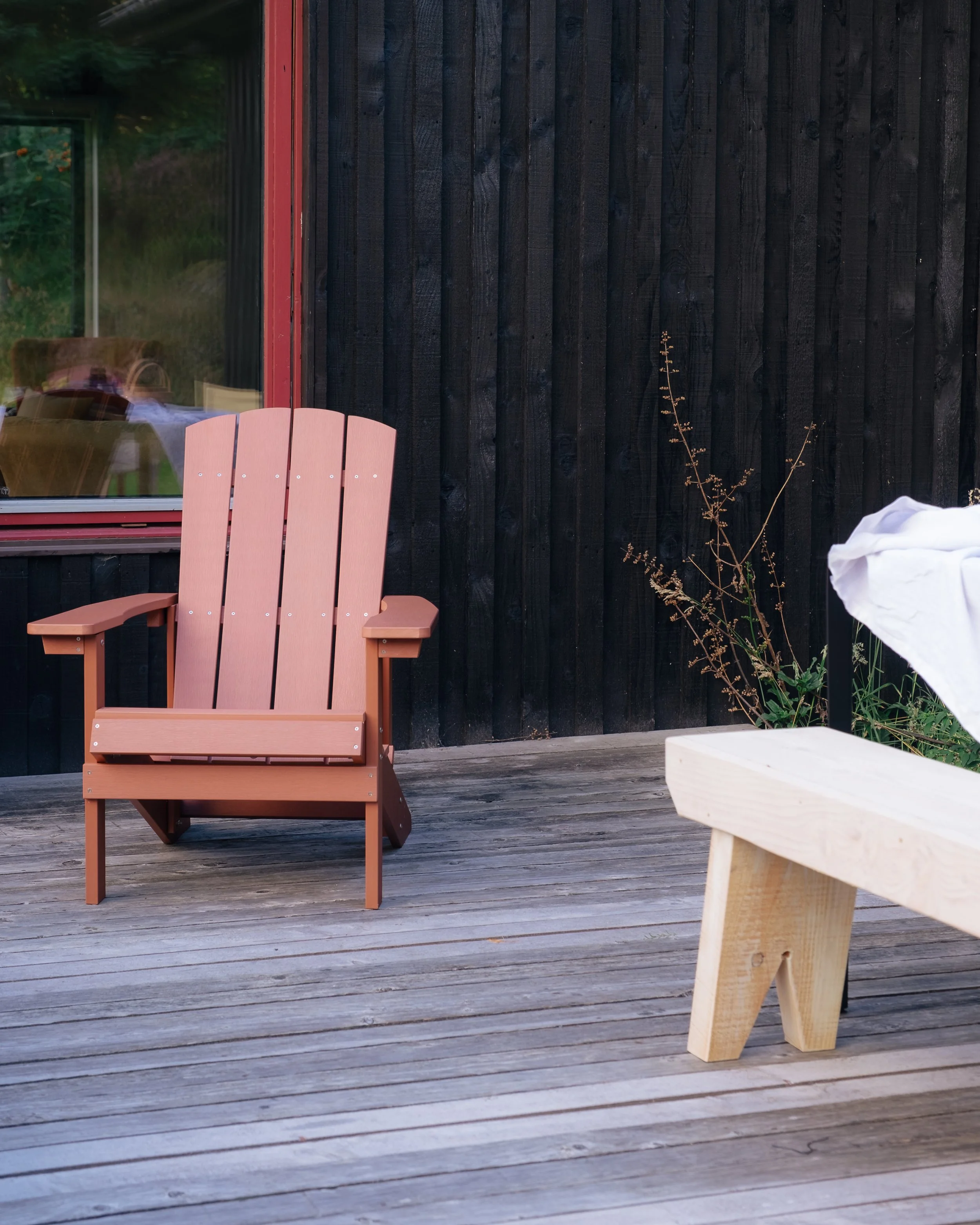 A patio with a pink Adirondack chair, a small wooden bench, and some plants against a black wooden wall, with a window showing a reflection of the outside.