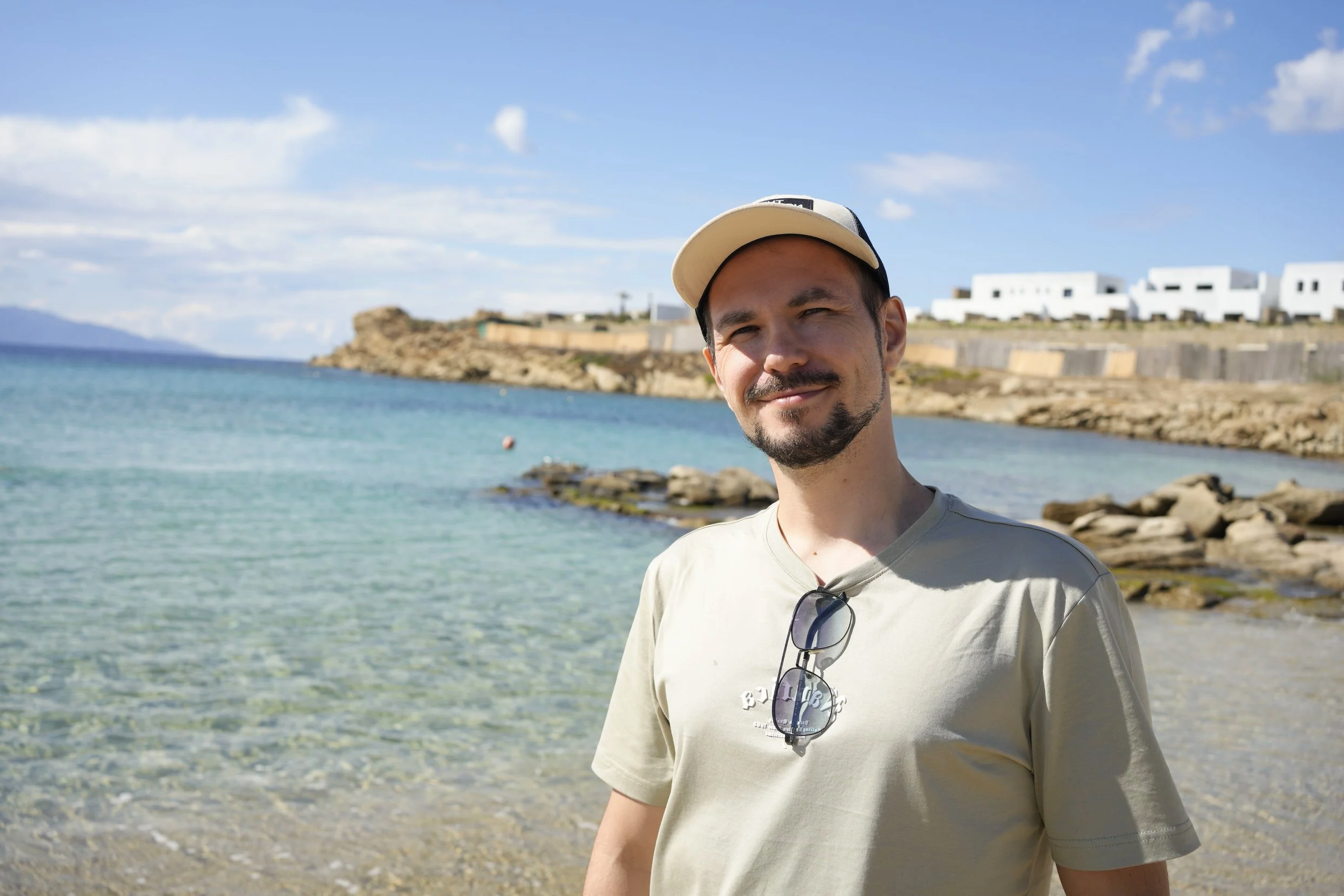 A young man with a beard smiling outdoors by the water on a sunny day, wearing a beige t-shirt, a beige cap, and sunglasses hanging from his shirt collar, with white buildings on a rocky shoreline in the background.