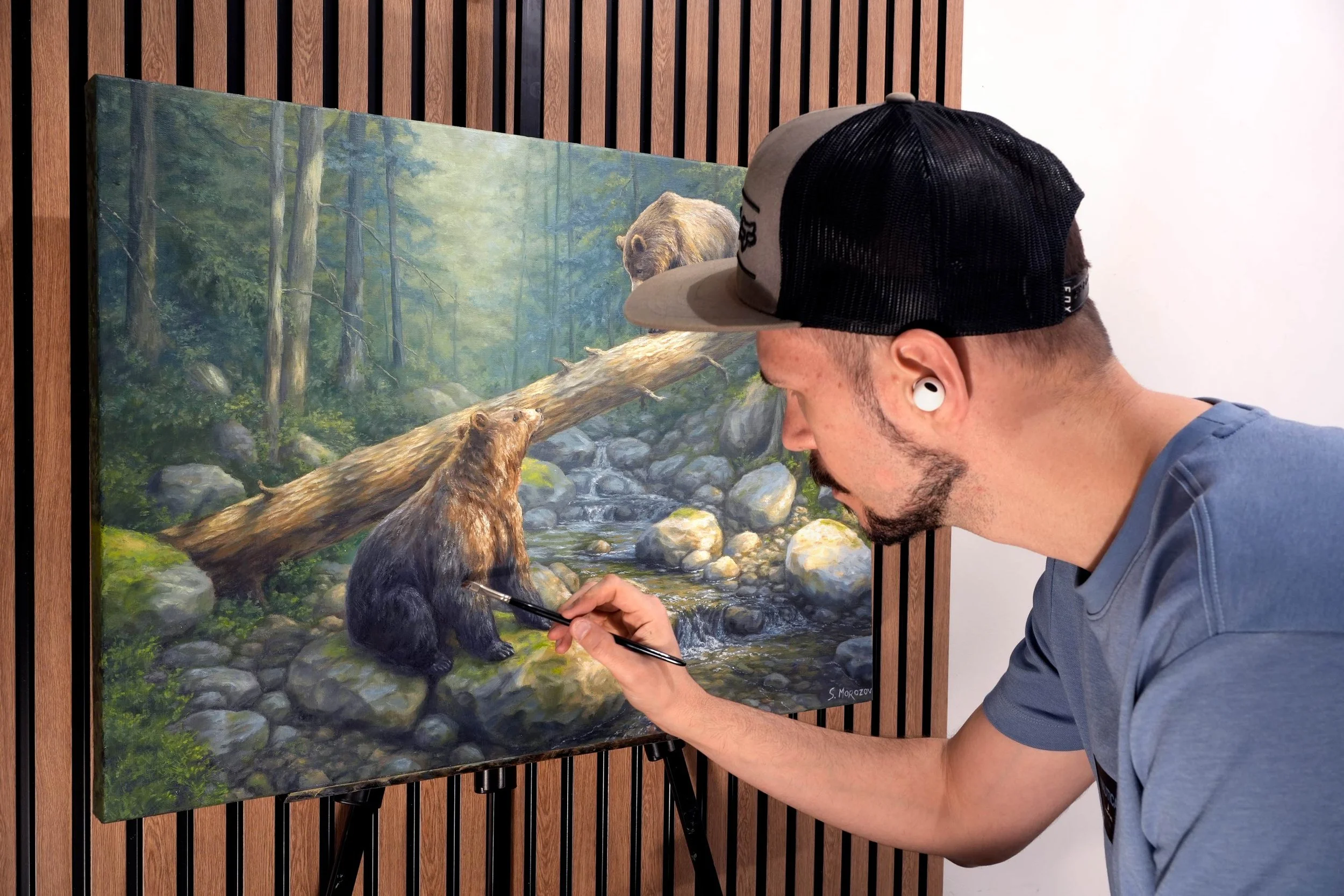 An artist painting a realistic nature scene with bears in a forest stream on a canvas. The painter wears a baseball cap and wireless earbuds.